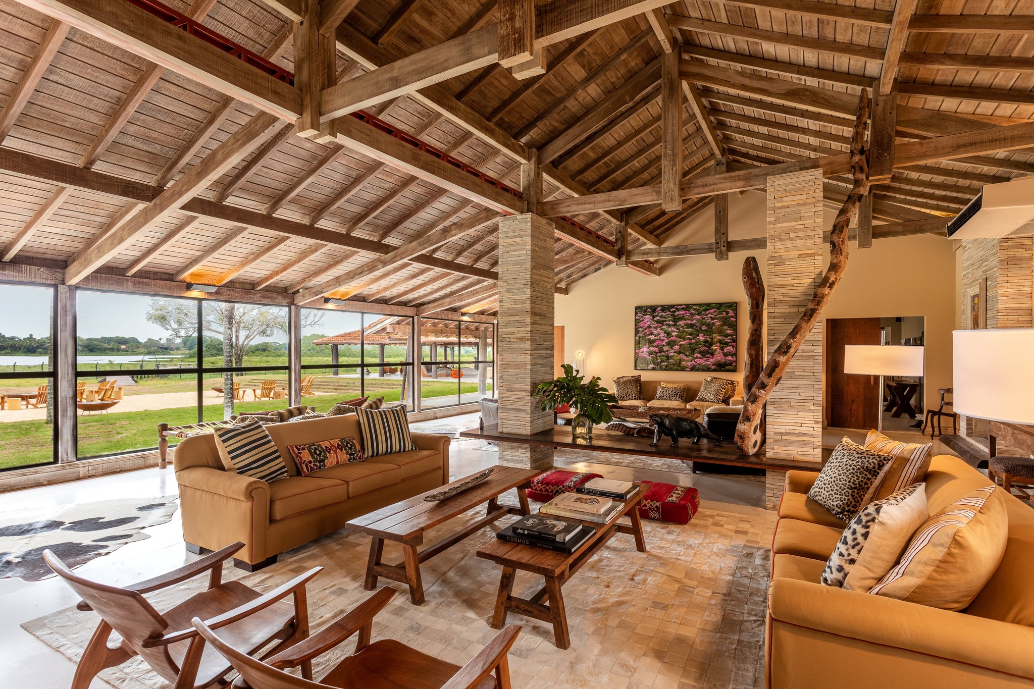 Rustic grandeur meets open sky — the lodge's soaring rafters and amber sofas invite you to unwind between safaris. Soaring timber-beam ceiling and ochre sofas in the open-plan lodge living room of a pantanal luxury safari property, with floor-to-ceiling glass walls framing green wetland views beyond
