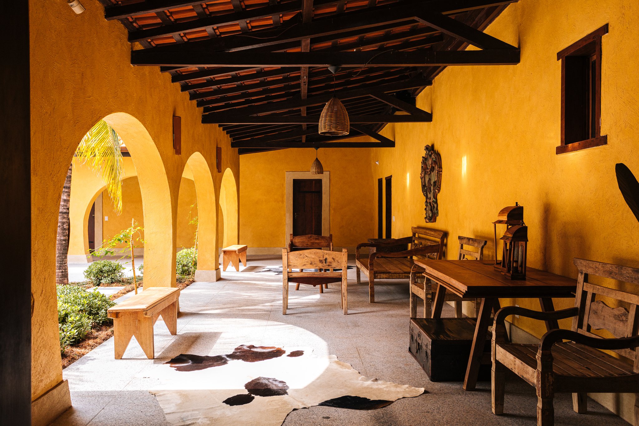 Handcrafted timber, arched ochre walls, and afternoon light — the lodge's veranda sets the tone from the moment you arrive. Sunlit covered veranda of a pantanal luxury safari lodge, with golden ochre arched walls, dark timber beam ceiling, rustic wooden benches and dining table, wicker pendant lights, and a cowhide rug on the stone floor
