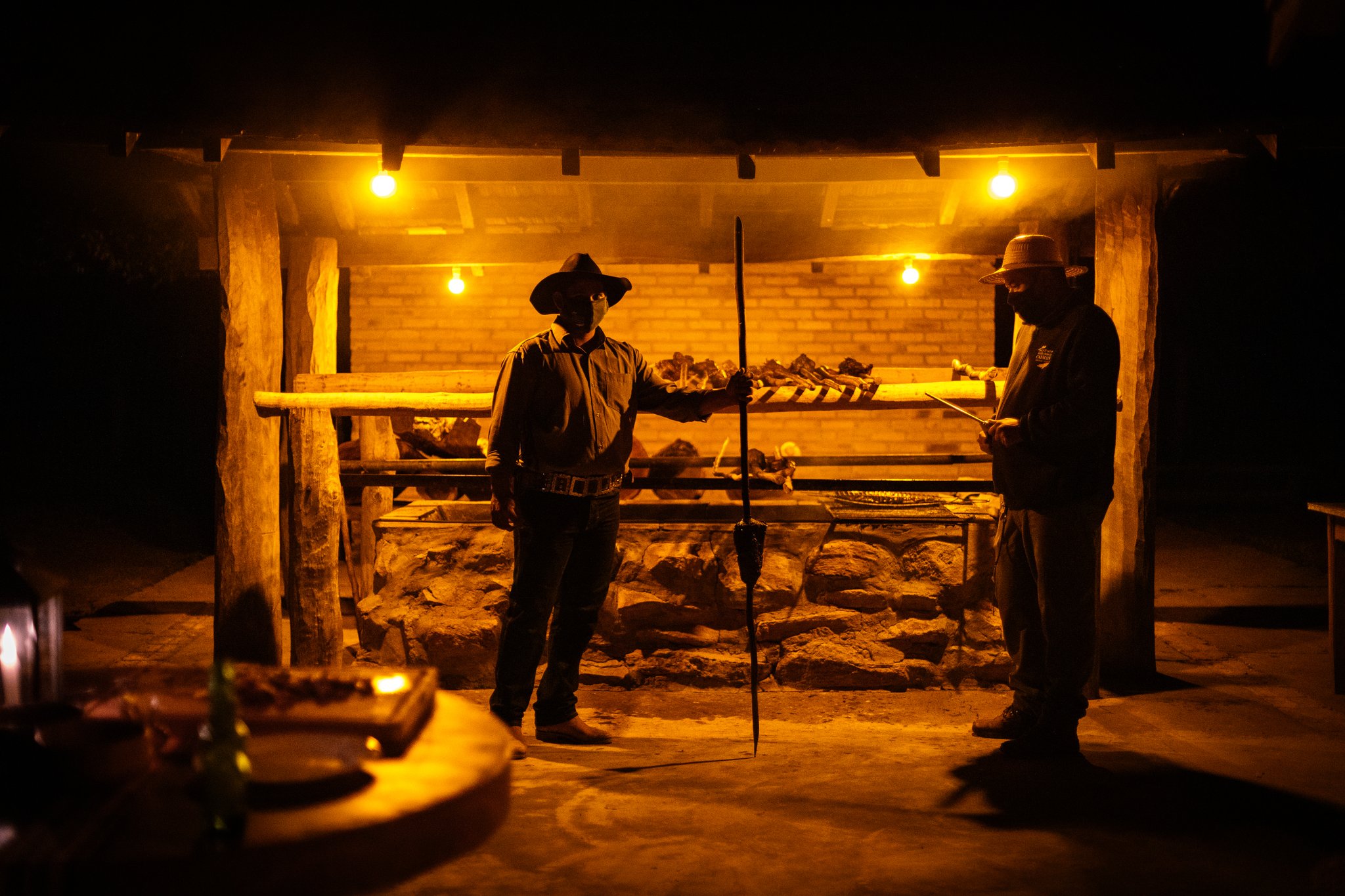 Two hats, one fire, a whole spit — dinner earned the slow way. Two gauchos in wide-brimmed hats tend a large open-fire wood-spit grill glowing amber at night during a pantanal luxury safari dinner at Caiman