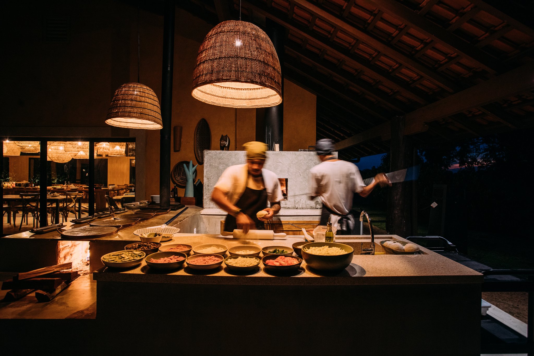 Bowls of fresh ingredients, a wood-fired oven, two chefs in full flight — dinner is well underway. Two chefs in motion at an open-air wood-fired kitchen station during a pantanal luxury safari dinner, with bowls of fresh ingredients lit by warm wicker pendant lamps