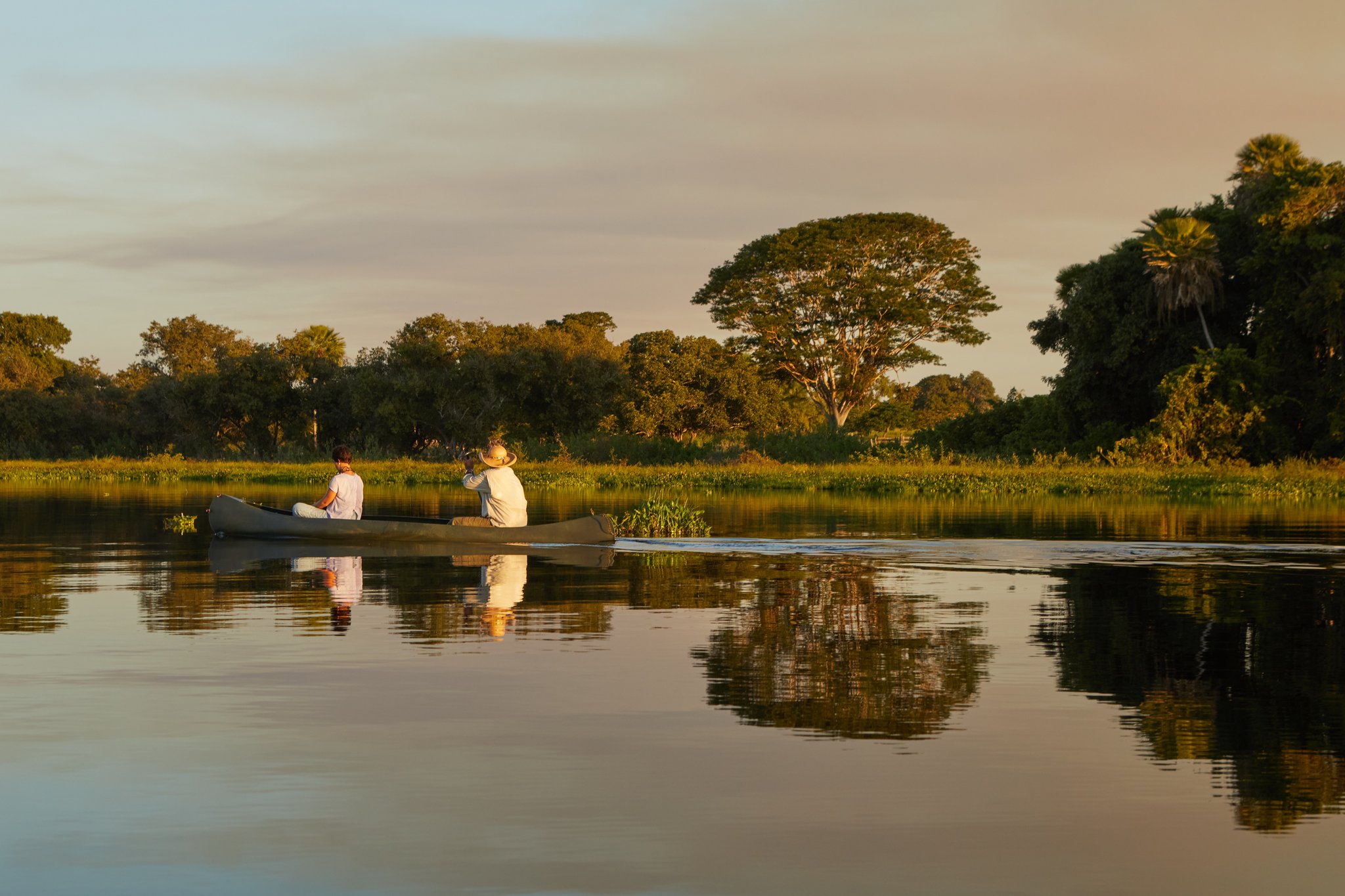 Paddling softly into the last light of the day. Two guests drifting in a canoe on glassy water at golden hour during a pantanal luxury safari, surrounded by lush wetland vegetation and large trees reflected in the still river