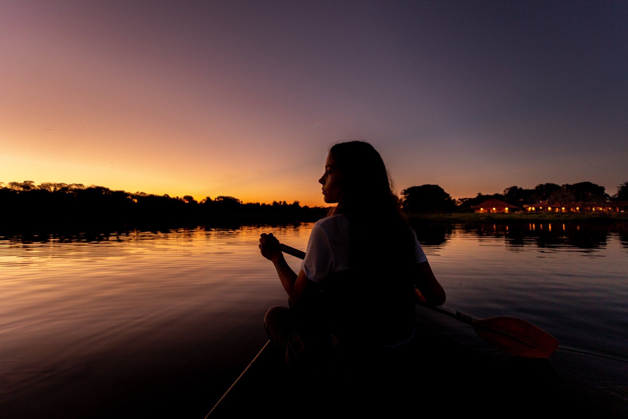 Paddle in hand, the whole sky turning amber. Silhouette of a woman holding a canoe paddle on a glassy river during a pantanal luxury safari, with a vivid orange and purple sunset reflected on the water and lodge lights glowing on the far bank