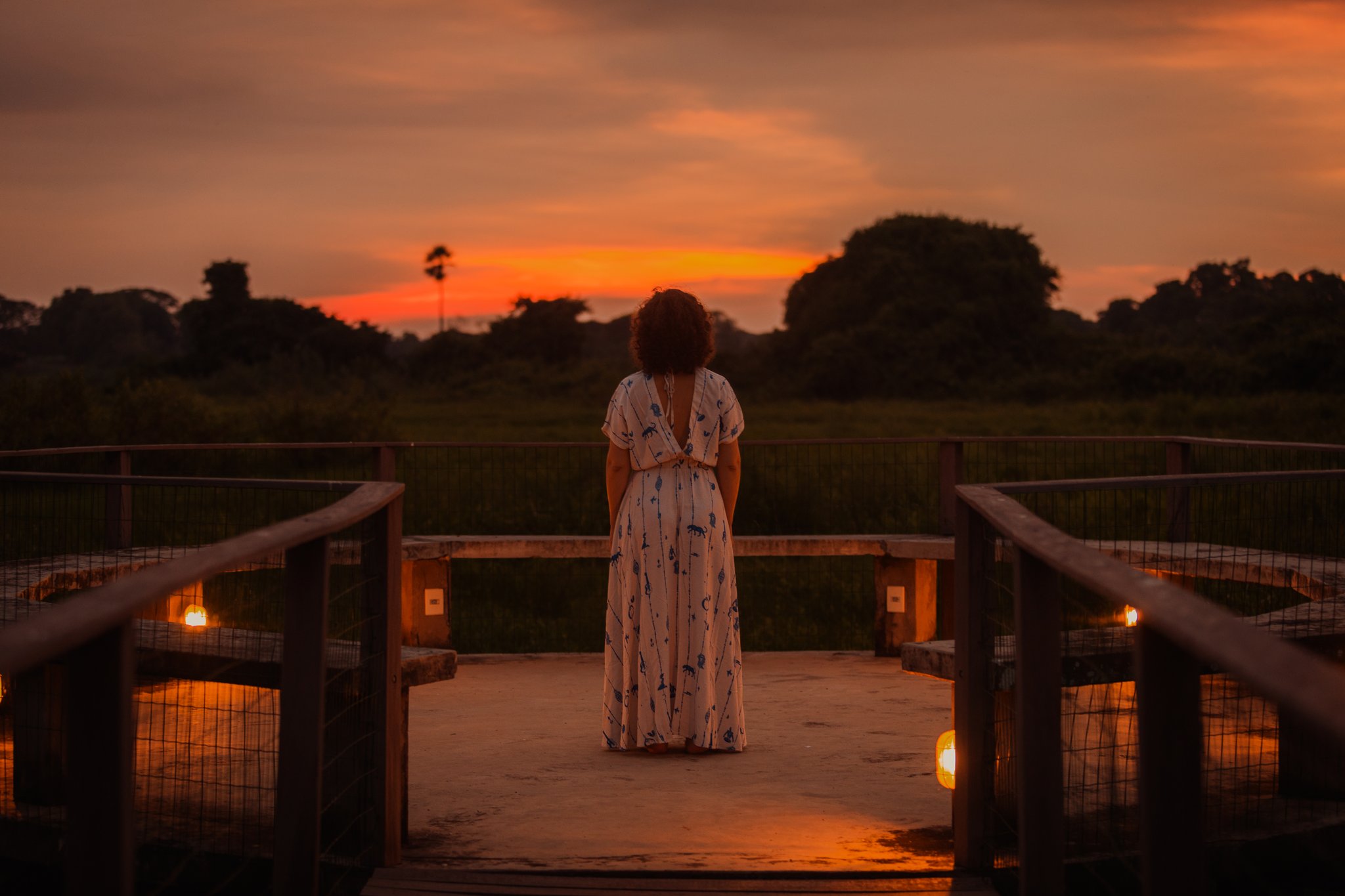 Still skies, burning horizons — evenings here belong entirely to you. A woman in a floral maxi dress stands on a candlelit wooden deck, watching a blazing orange sunset over open wetland flats on a pantanal luxury safari