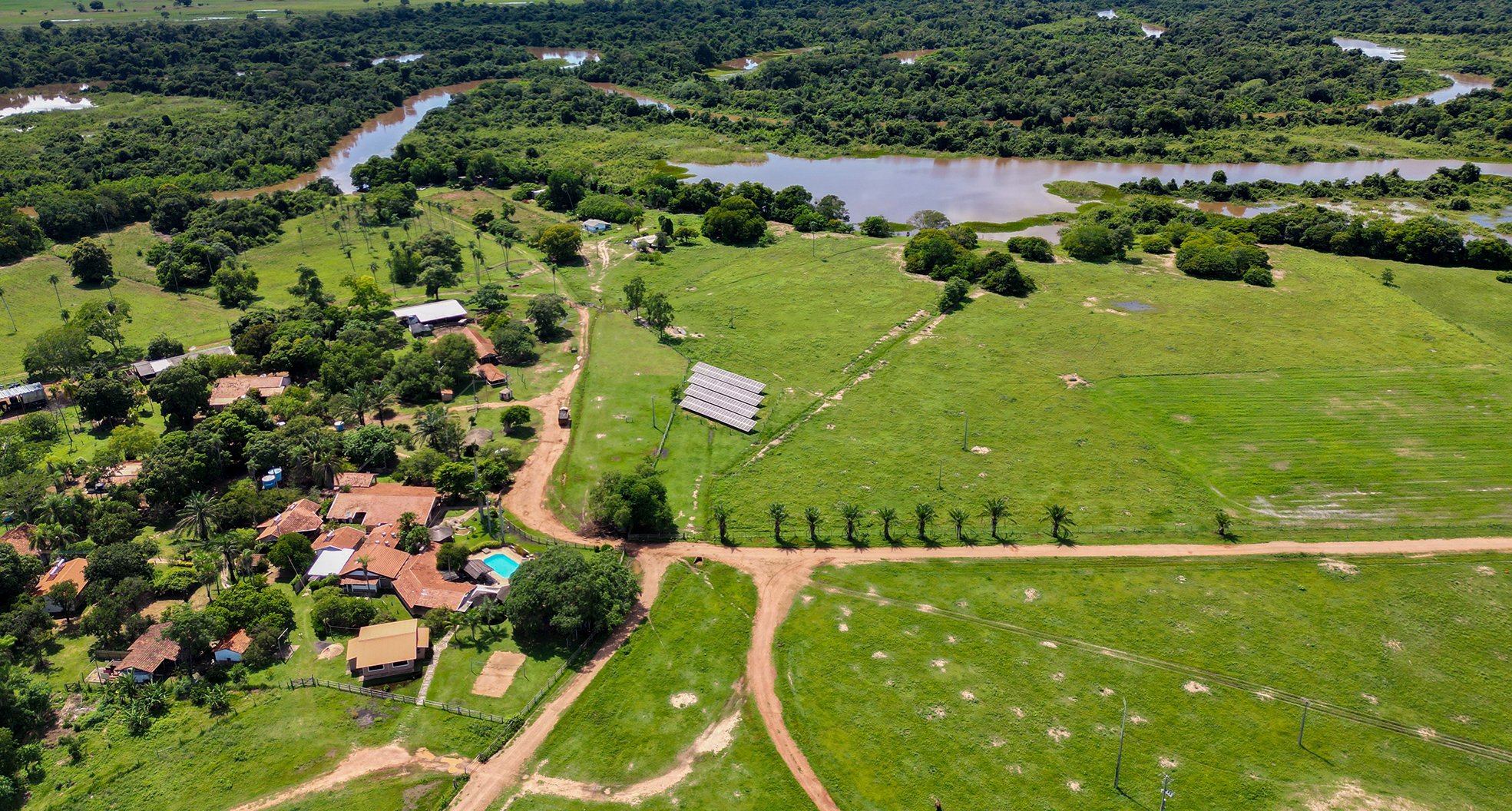 Solar-powered and river-kissed — an ecolodge nestled between wetland pastures and the wild horizon. Aerial view of a Pantanal ecolodge on a pantanal tour from Campo Grande, with solar panels, red-roofed buildings, a swimming pool, dirt tracks, green pastures, and a winding river visible in the background