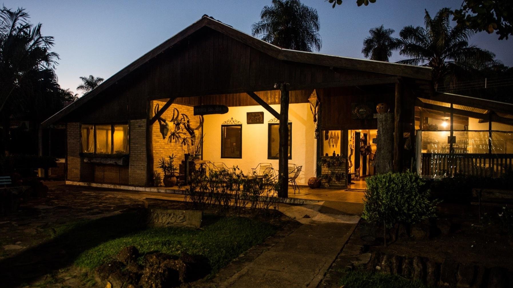Amber light spills from the veranda as the Pantanal night begins. A warmly lit rustic ecolodge with a wooden gabled roof and open veranda glowing with amber light at dusk, surrounded by palm trees and tropical gardens, on a pantanal tour from campo grande