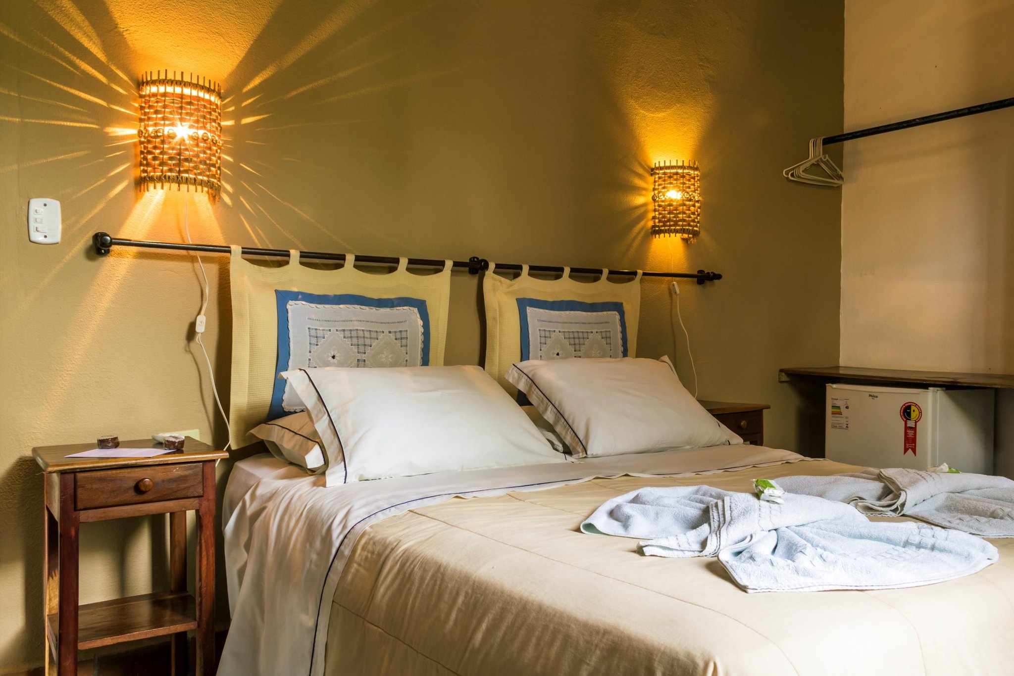 Soft lamplight, fresh robes, and a comfortable bed waiting after a day in the wetlands. Cozy double room at a Pantanal tour from Campo Grande ecolodge, featuring warm woven wall sconces, cream bedding with decorative pillows, folded white towels and robes on the bed, a wooden nightstand, and a mini-fridge