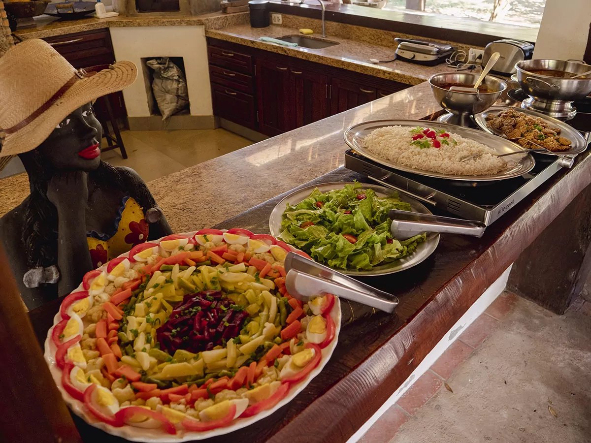 Home-cooked abundance, ready the moment you return from the wetlands. A colourful buffet spread at a Pantanal tour from Campo Grande ecolodge, featuring a decorated vegetable and egg salad platter, fresh green salad, rice garnished with red peppers, and fried dishes on a granite counter beside a traditional ceramic figurine