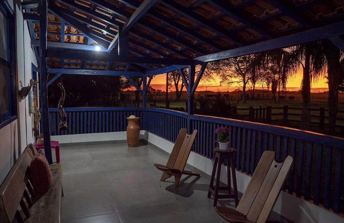 A covered veranda with blue-painted wooden railings and rustic chairs overlooks a wide open landscape glowing with a deep orange sunset on a pantanal tour from campo grande