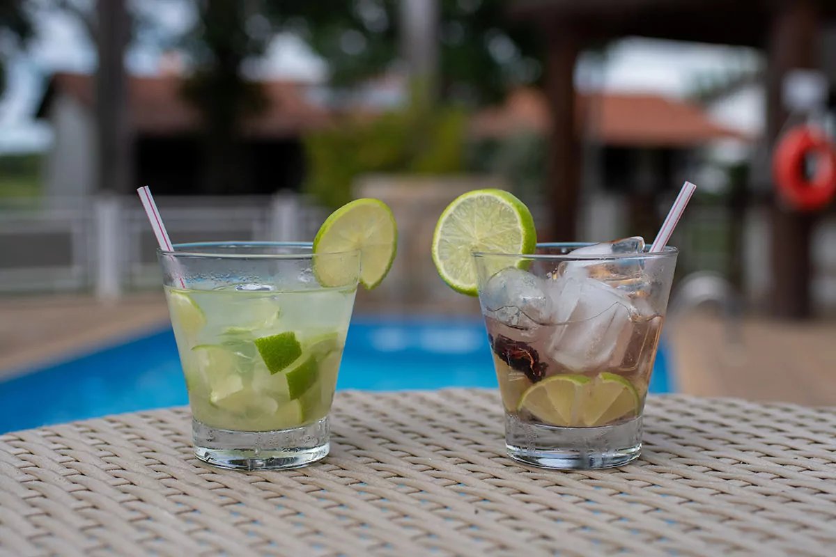 Ice-cold caipirinhas — the perfect reward after a day in the wetlands. Two lime-garnished cocktails with ice and straws on a wicker table beside a blue swimming pool at a Pantanal tour from Campo Grande ecolodge