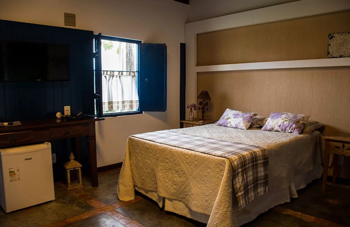 A cozy double room on a pantanal tour from campo grande, featuring a quilted bedspread with floral pillows, blue wooden shutters open to garden greenery, a wooden desk with flat-screen TV, and a mini-fridge on terracotta tile floors
