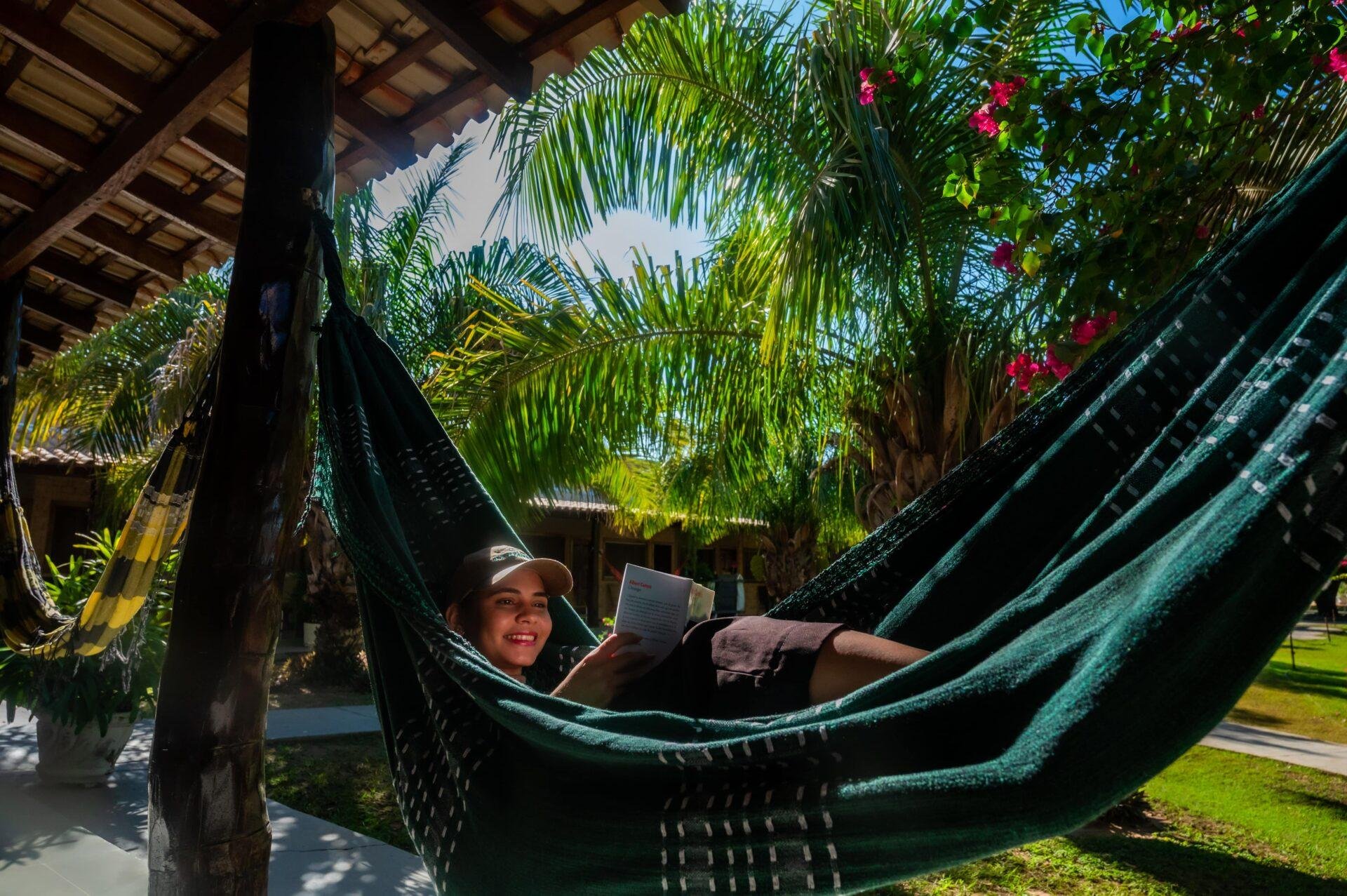 Book in hand, nowhere else to be. A smiling traveller reads a book while resting in a dark green hammock on a shaded lodge veranda, surrounded by palm fronds and pink bougainvillea, during a Pantanal Tour from Cuiabá