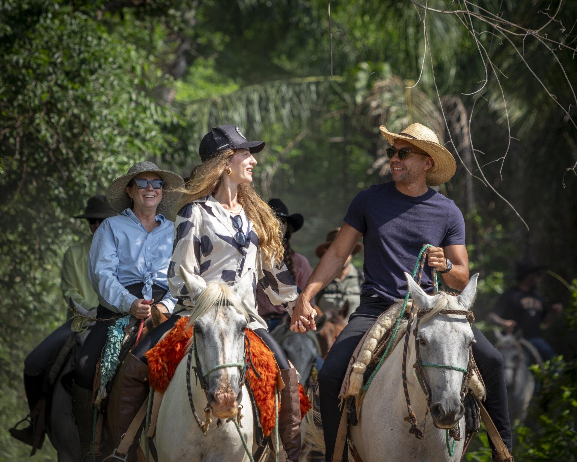Laughing, hand-in-hand, deep in the wetland trails. A group of smiling riders on grey horses moves through a lush green corridor on a Pantanal Tour from Cuiabá, a couple in the foreground holding hands in the saddle