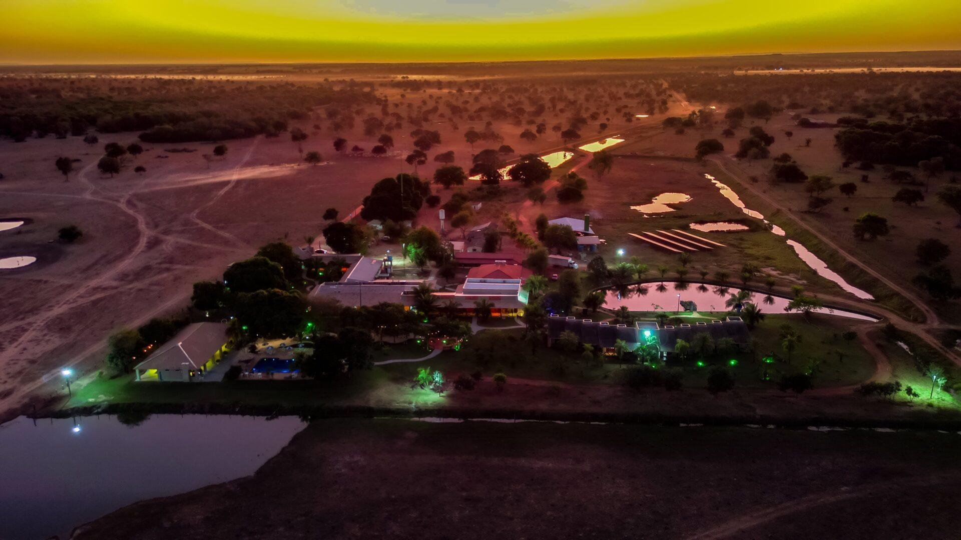 Glowing under a golden dusk sky, the lodge sits like a lit lantern in the vast wetland wilderness. Aerial drone view of an illuminated Pantanal lodge at dusk, surrounded by wetland ponds and open savanna scrubland stretching to the horizon under a vivid yellow-orange sky — a base for a Pantanal Tour from Cuiabá