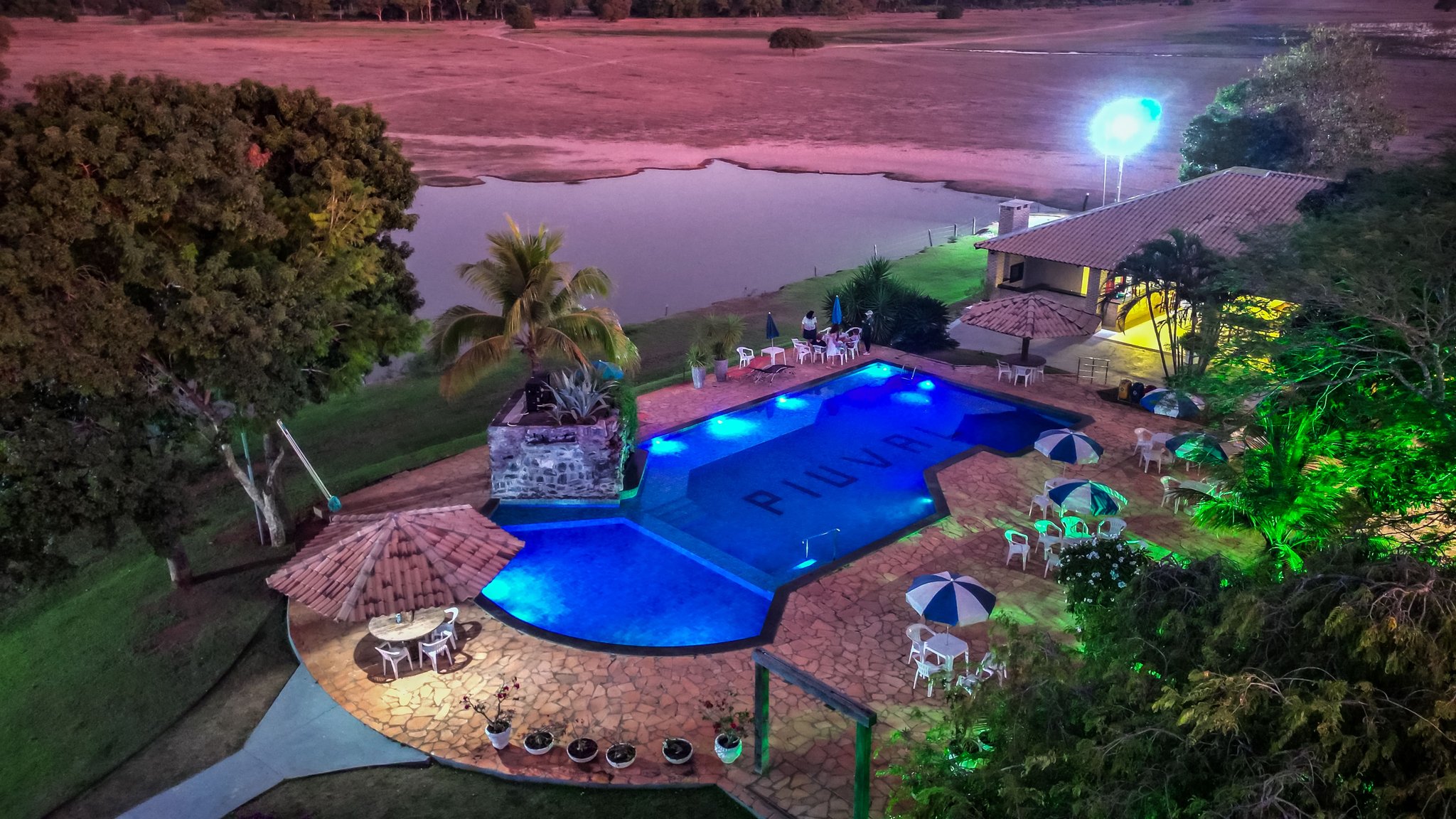Blue-lit pool, open skies, and a wild wetland just steps away. Aerial night view of an illuminated swimming pool at a Pantanal Tour from Cuiabá lodge, with patio umbrellas, a stone water feature, and a natural lake stretching across open savanna in the background