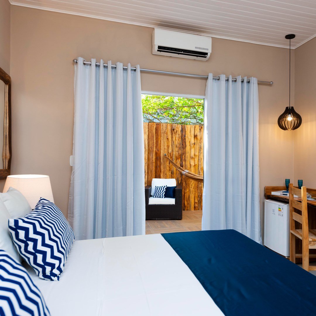 Rest well — your private terrace and hammock are just a curtain's width away. Air-conditioned double room on a Pantanal Tour from Cuiabá, with navy chevron pillows, white curtains, and a private wooden terrace with hammock and lounge chair visible through the open sliding door