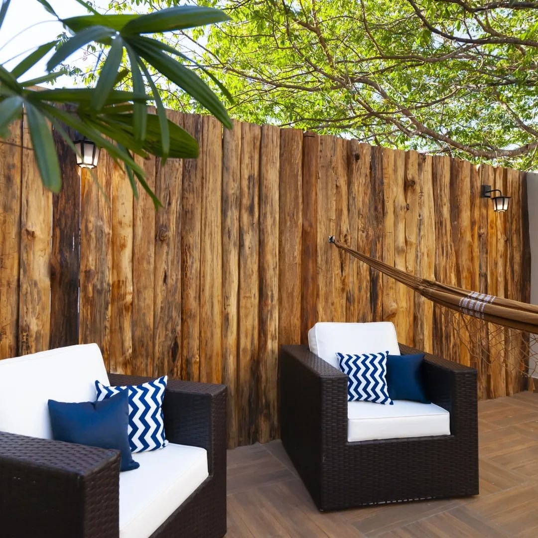 Rattan chairs, a swaying hammock, and tree shade overhead — unwind between wildlife adventures. Shaded terrace at a Pantanal Tour from Cuiabá lodge, with dark rattan armchairs, white cushions, blue chevron pillows, a hammock, and rustic timber walls beneath green tree canopy