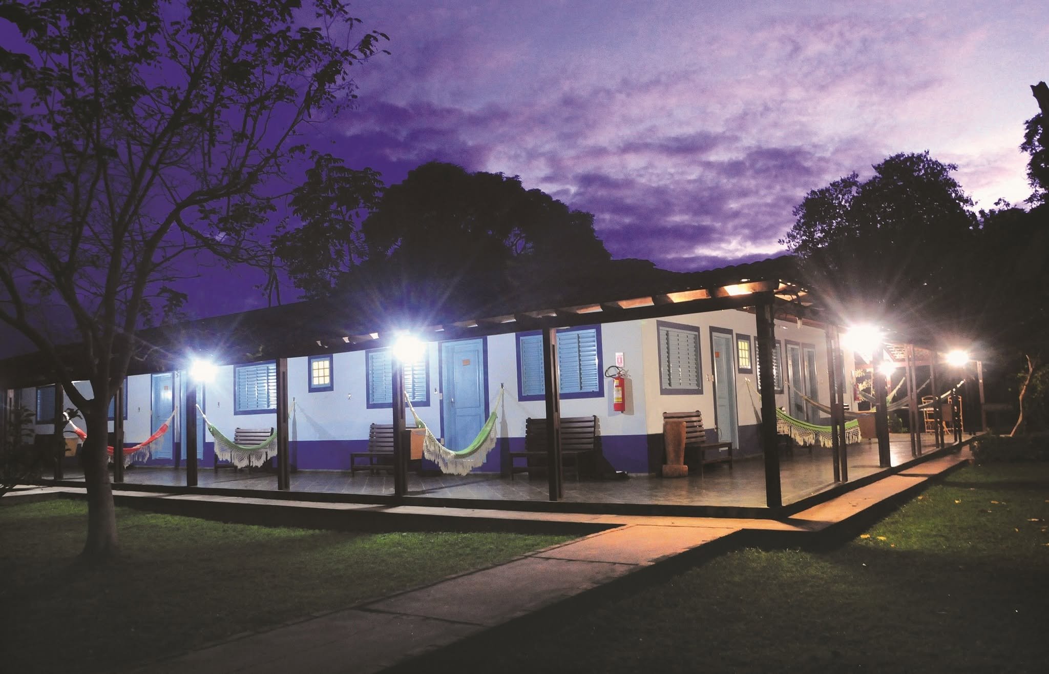 Hammocks ready, purple sky overhead — the night begins. A low white lodge building with colorful hammocks strung along a covered veranda, glowing under bright spotlights against a deep purple twilight sky on a Pantanal Tour from Cuiabá