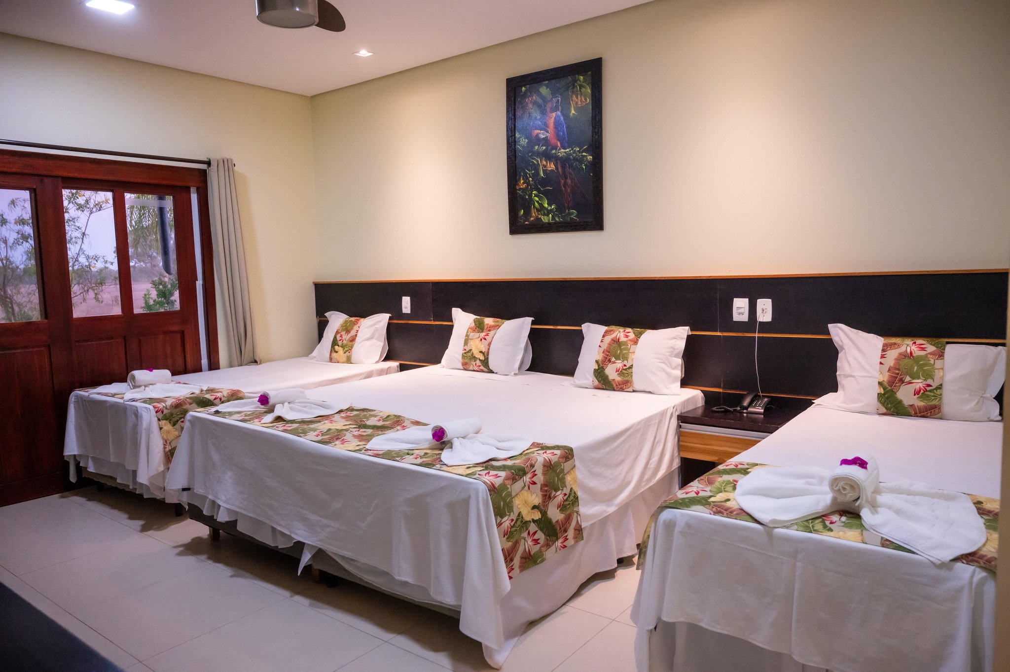 Three beds turned down, orchids on the pillows — your comfortable base for every early morning safari. A triple-occupancy lodge room on a Pantanal Tour from Cuiabá, with three neatly made beds featuring tropical floral runners, rolled towels with purple orchid accents, and a framed macaw painting above the headboard