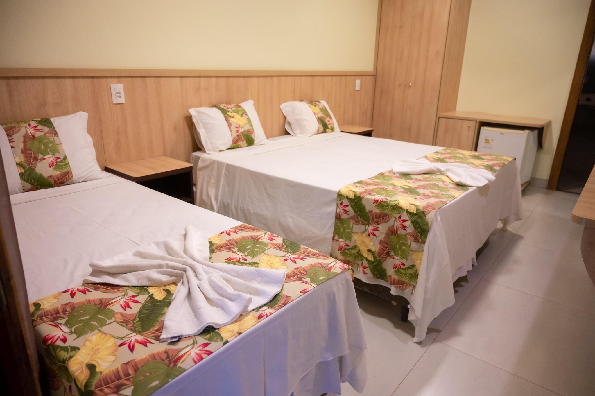 Fresh linens and tropical prints — rest well before the next day's adventure. Three neatly made beds with tropical floral bedspreads and folded white towels in a clean, wood-panelled guest room on a Pantanal Tour from Cuiabá