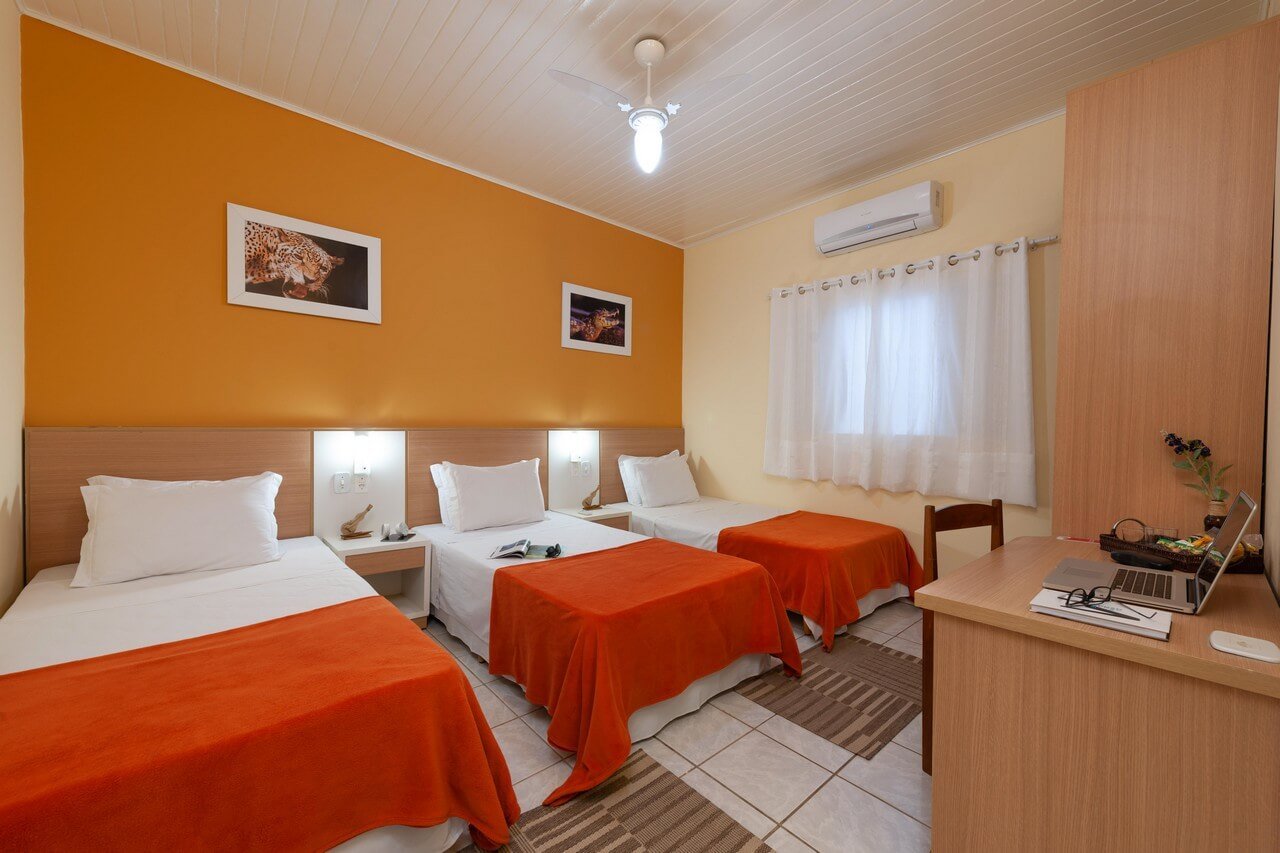 Orange walls, wildlife prints on every side — comfortable beds waiting after a full day in the wetlands. A bright triple room at a Pantanal Tour from Cuiabá lodge, featuring three single beds with orange throws, a warm amber accent wall adorned with wildlife prints, air conditioning, and a work desk with a laptop