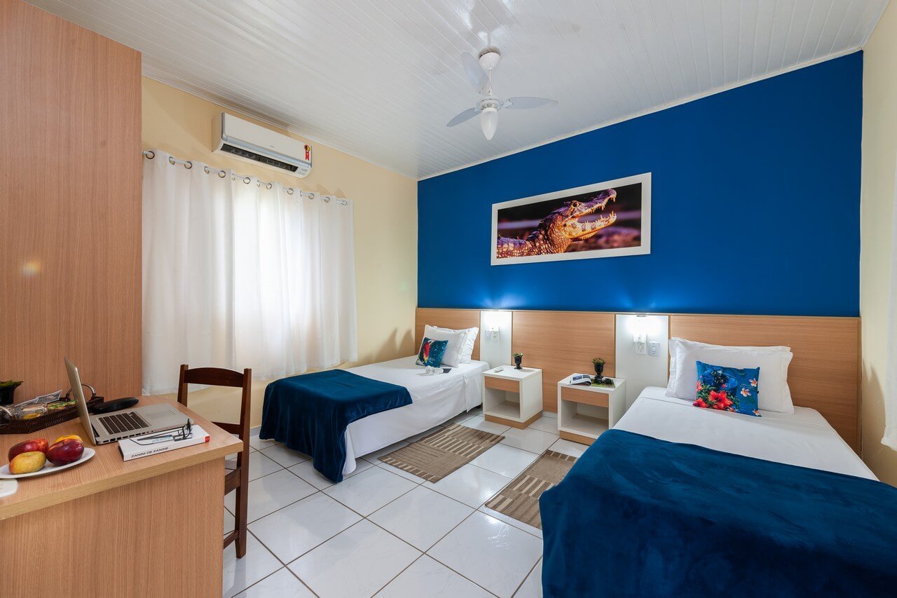 Navy throws, a caiman on the wall — your base between adventures. Twin room at a Pantanal Tour from Cuiabá lodge, featuring two beds with navy throws, a bold blue accent wall, a framed caiman photograph, and a work desk with fresh fruit