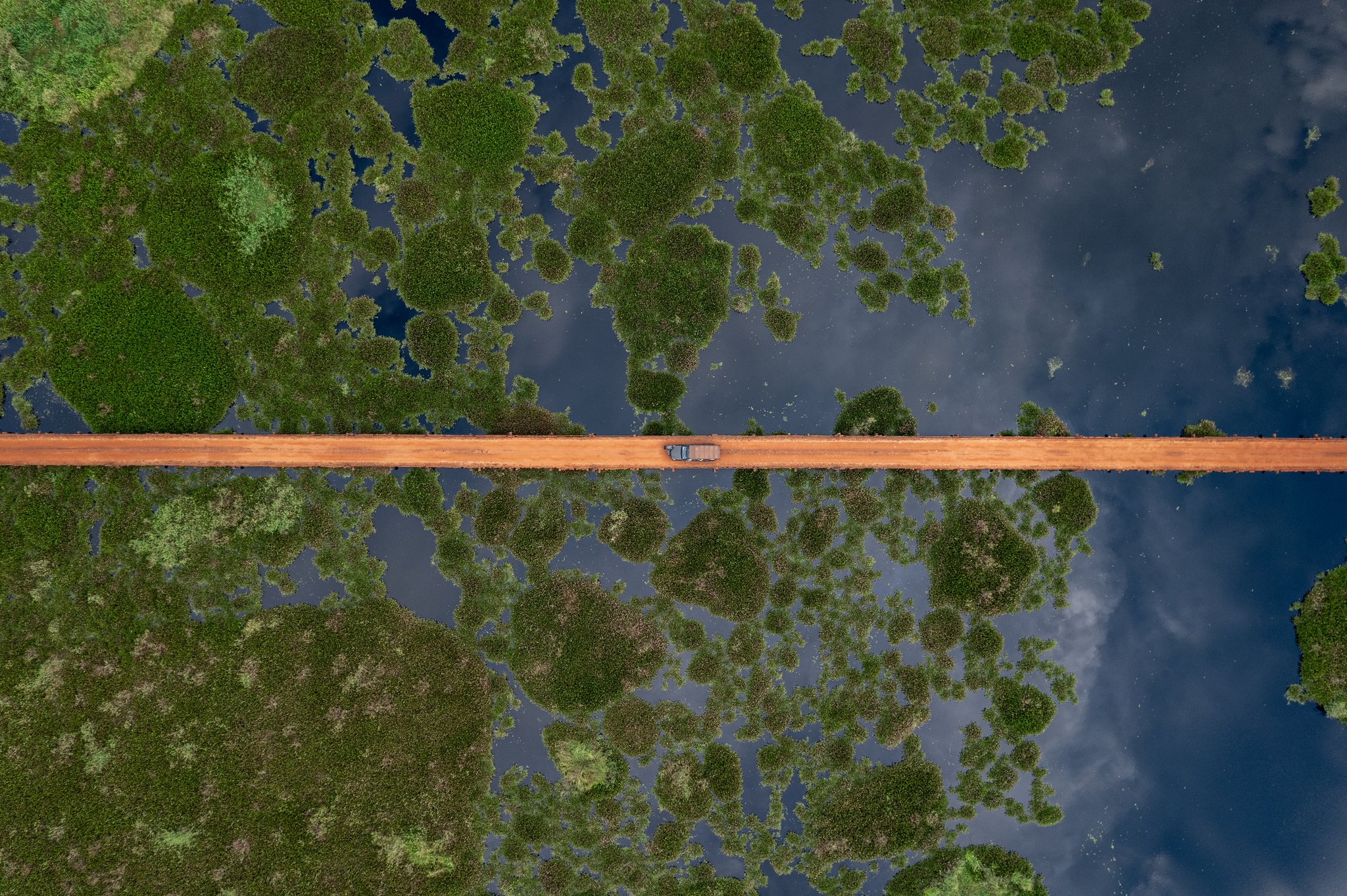 Vista aérea de uma estrada de terra vermelha cortando o Pantanal Sul, com um veículo pequeno ao centro cercado por ilhas de vegetação e águas espelhadas do período de cheia