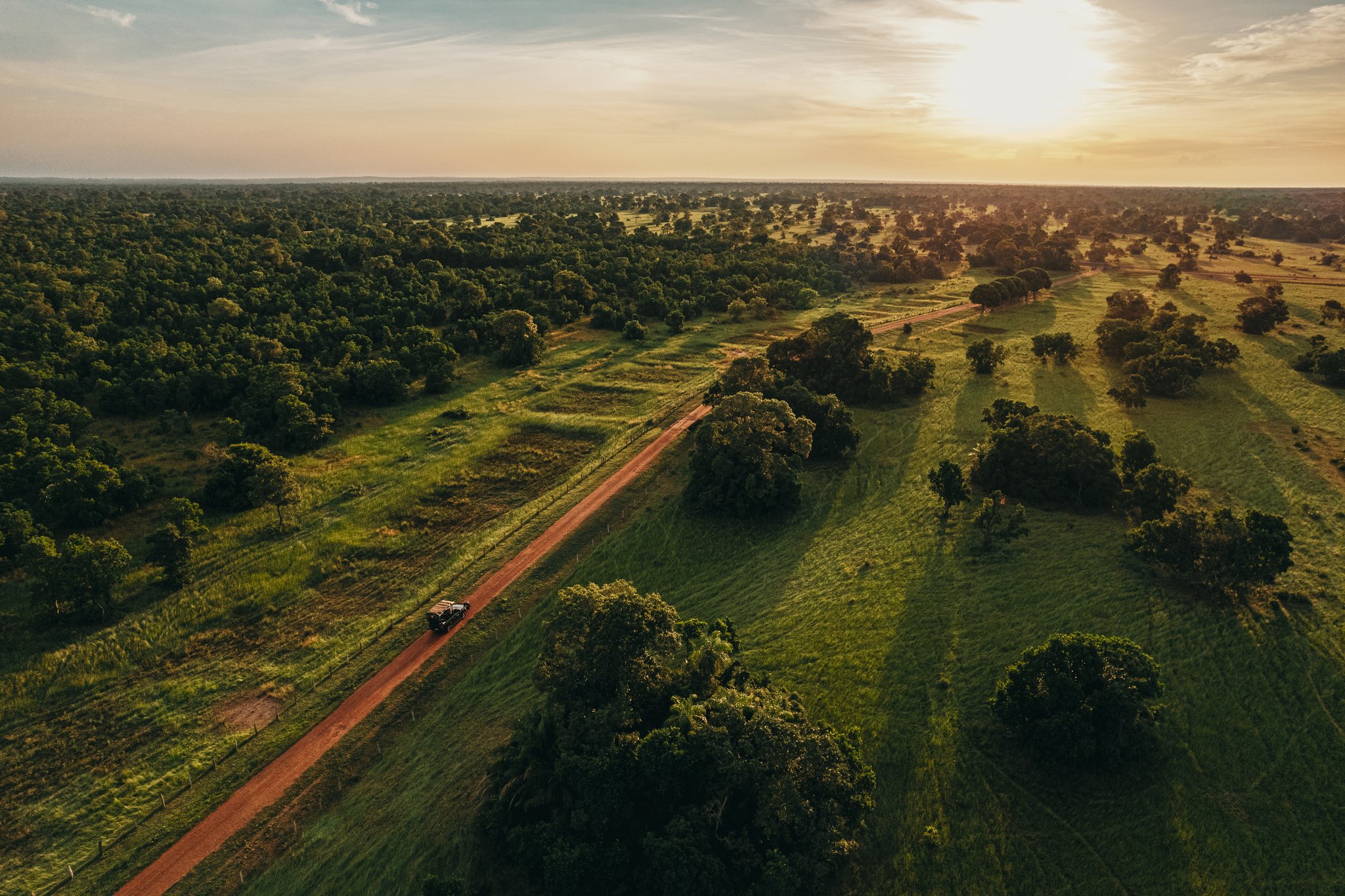 Vista aérea de um veículo 4x4 percorrendo uma estrada de terra vermelha pelo vasto Pantanal Sul ao pôr do sol, rodeado de cerrado e campos abertos