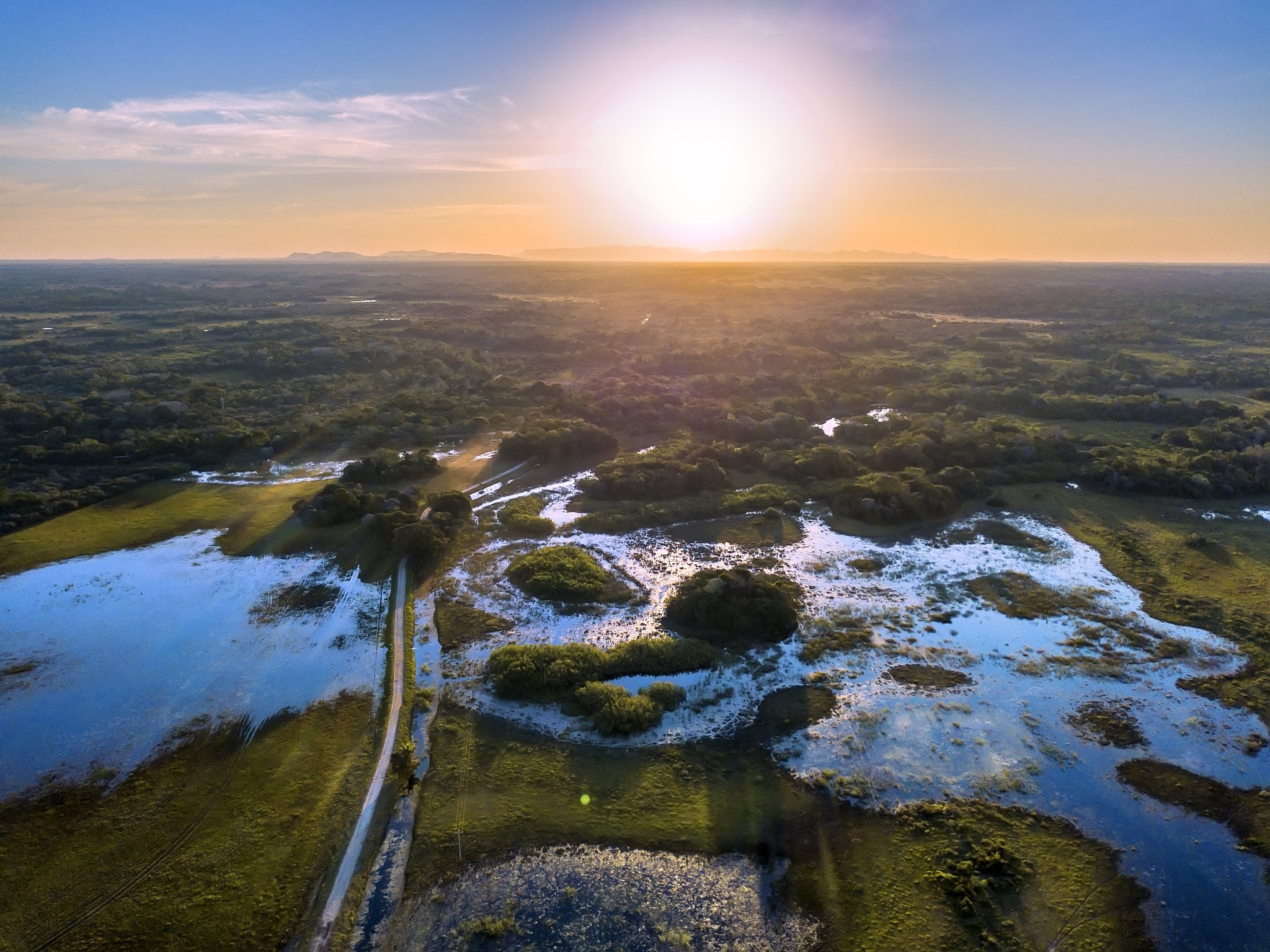 Aerial view of the Pantanal tours Brazil landscape at golden sunset, showing vast flooded wetlands, green vegetation islands, and a dirt track cutting through the waterlogged plains