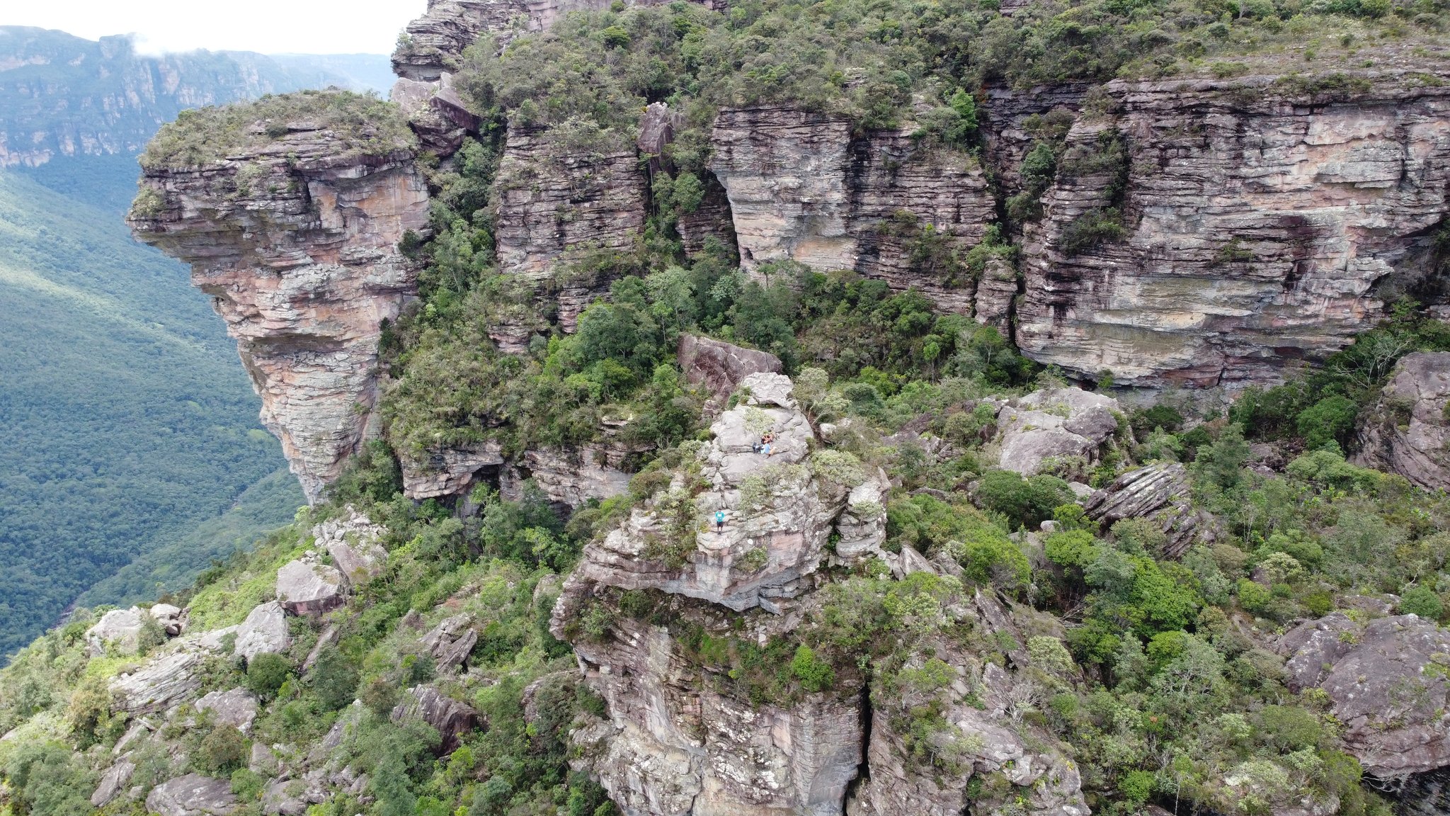 Aerial view of layered sandstone cliffs and dense green vegetation during a Pati Valley hike, with tiny hikers visible on a rocky outcrop far below