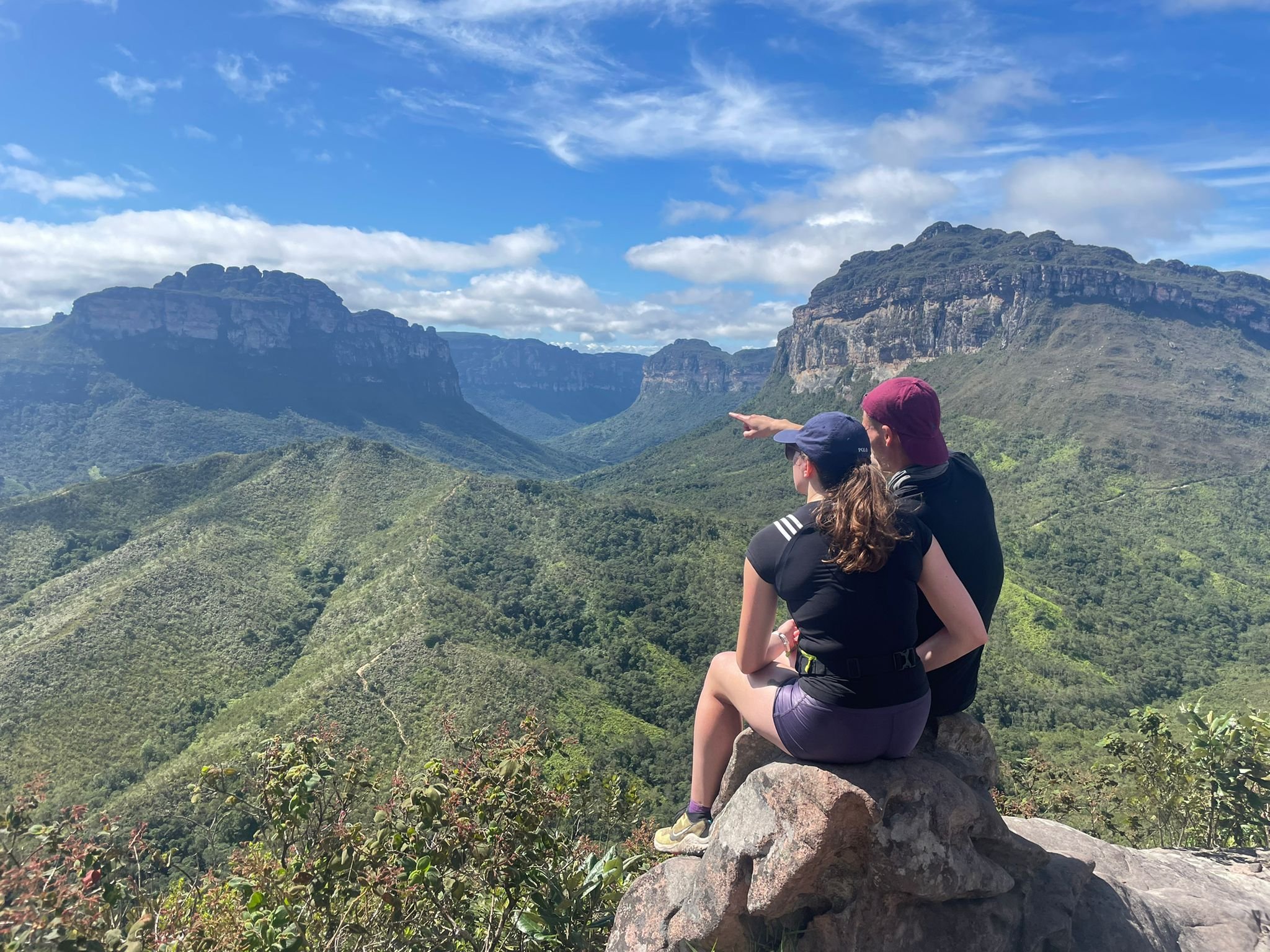 Pointing out what words can't quite capture. Photo: Lucas Neves Two hikers sit on a rocky outcrop on the Pati Valley hike, one pointing toward the vast green canyon flanked by towering sandstone plateaus under a blue sky
