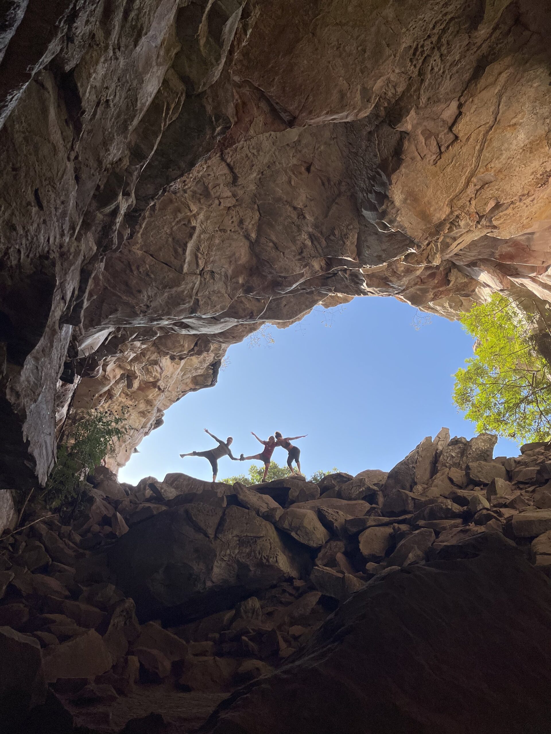 Balance found inside ancient rock — the cave at Morro do Castelo puts everything in perspective. Two hikers striking balance poses on boulders framed by a massive sandstone cave arch on the Pati Valley hike, blue sky and green treetops visible through the opening