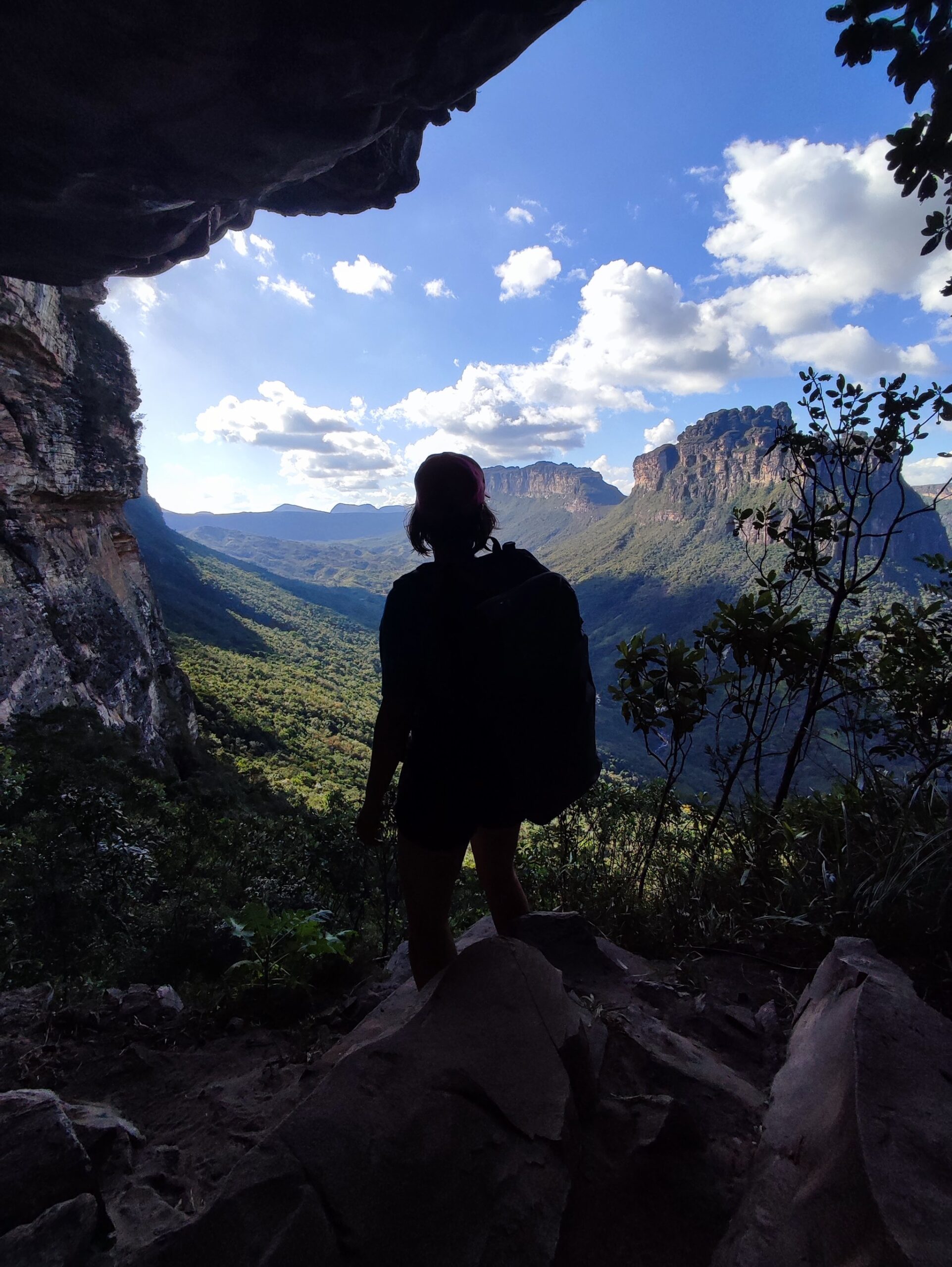 Framed by ancient rock, the whole valley opens up. A hiker's silhouette stands beneath a rocky overhang on the Pati Valley hike, gazing out over a sweeping green canyon and flat-topped sandstone escarpments under a blue sky