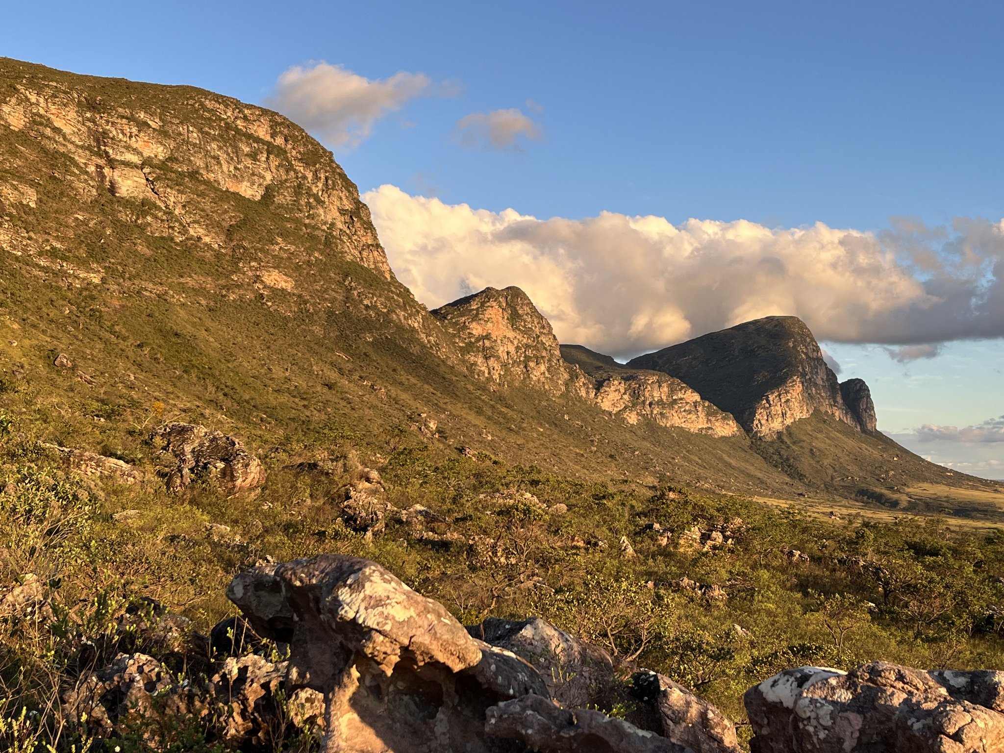 Warm golden light bathes the rocky escarpments and low scrubland of the Pati Valley hike, with billowing clouds rising behind layered sandstone cliffs