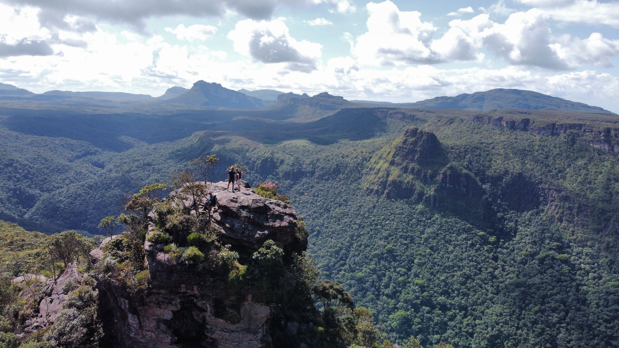 Tiny silhouettes, an ocean of canyon below — the scale of Chapada Diamantina earned one step at a time. Photo: Alex Hubin Two hikers stand on a mossy sandstone pinnacle during the Pati Valley hike, overlooking a vast canyon covered in dense green forest with layered tabletop mountains stretching to the horizon under a partly cloudy sky