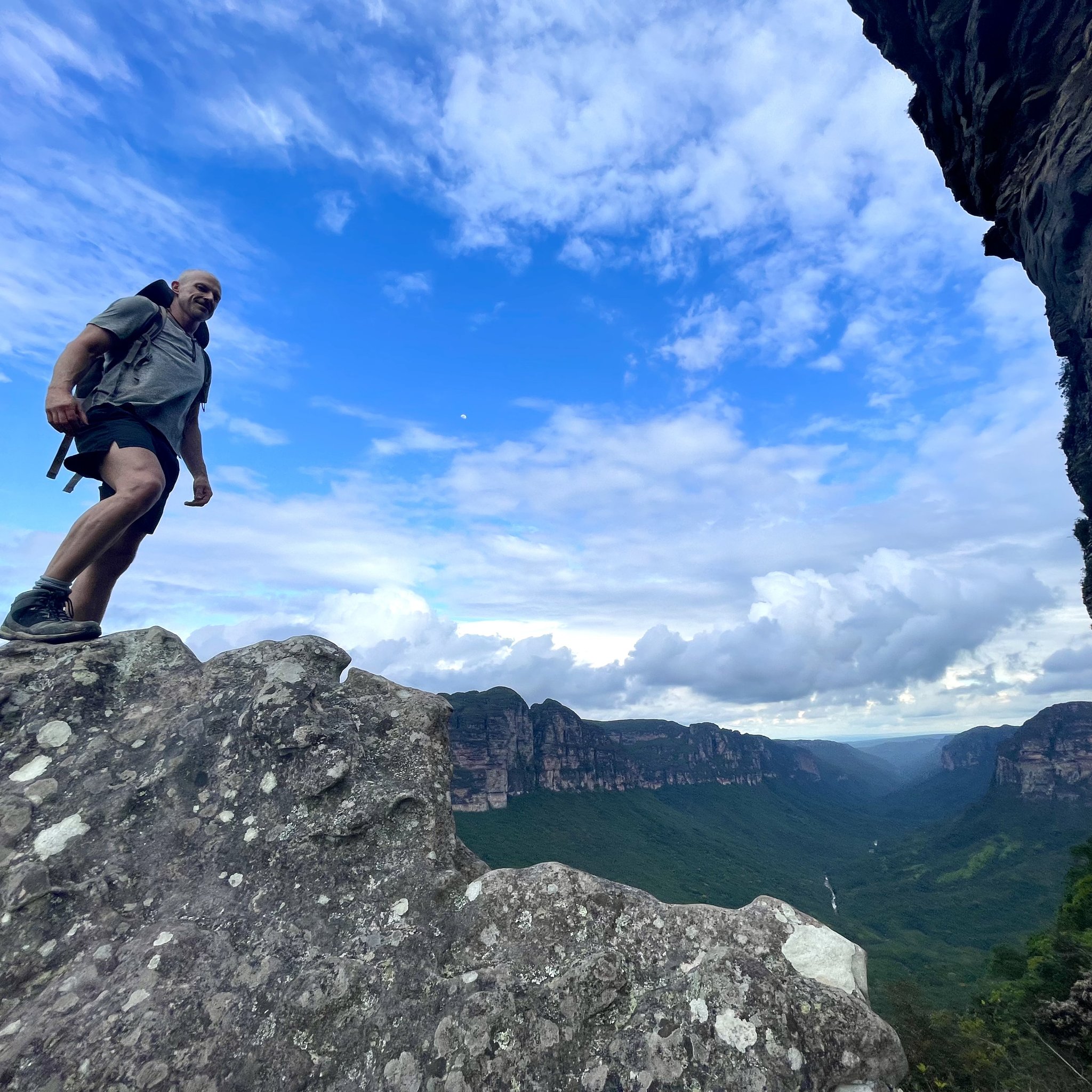 A hiker with a backpack stands on a rocky summit during the Pati Valley hike, with vast canyon walls and green valley stretching far below under a wide blue sky