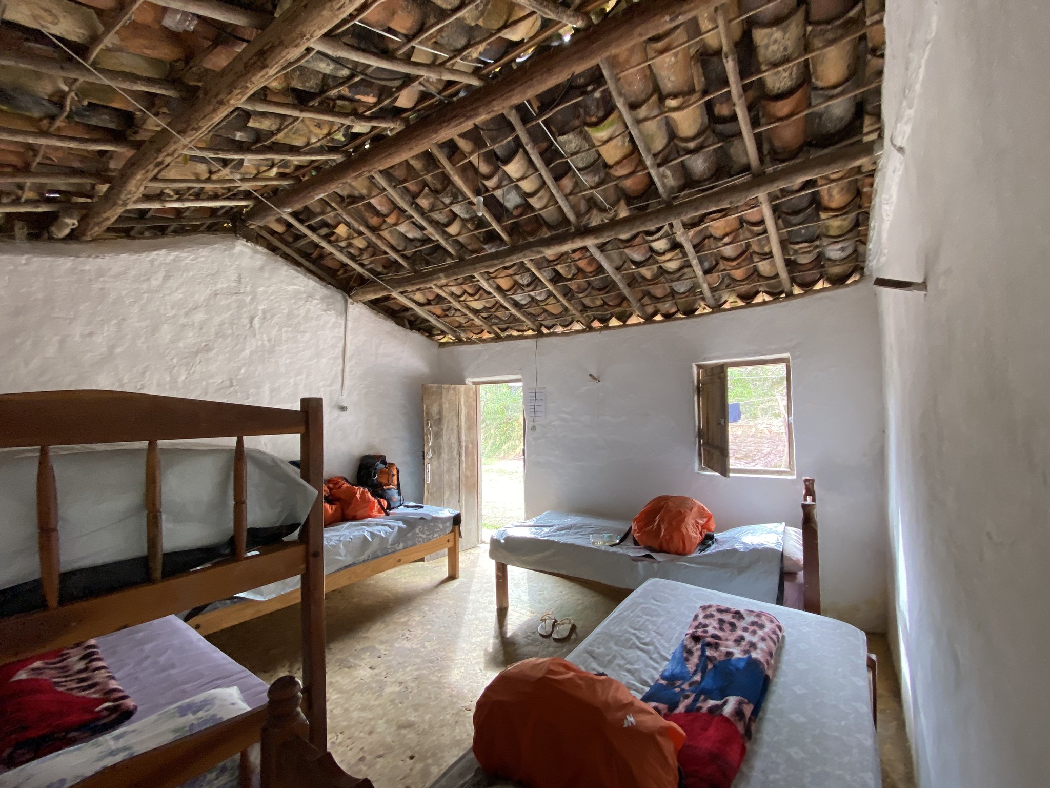 Rustic and real — terracotta tiles overhead, wooden bunks, and your gear waiting for tomorrow's trail. Simple shared dormitory room on the Pati Valley hike, with wooden bunk beds, white-washed walls, exposed clay-tile roof on timber beams, and trekkers' orange dry bags resting on single mattresses