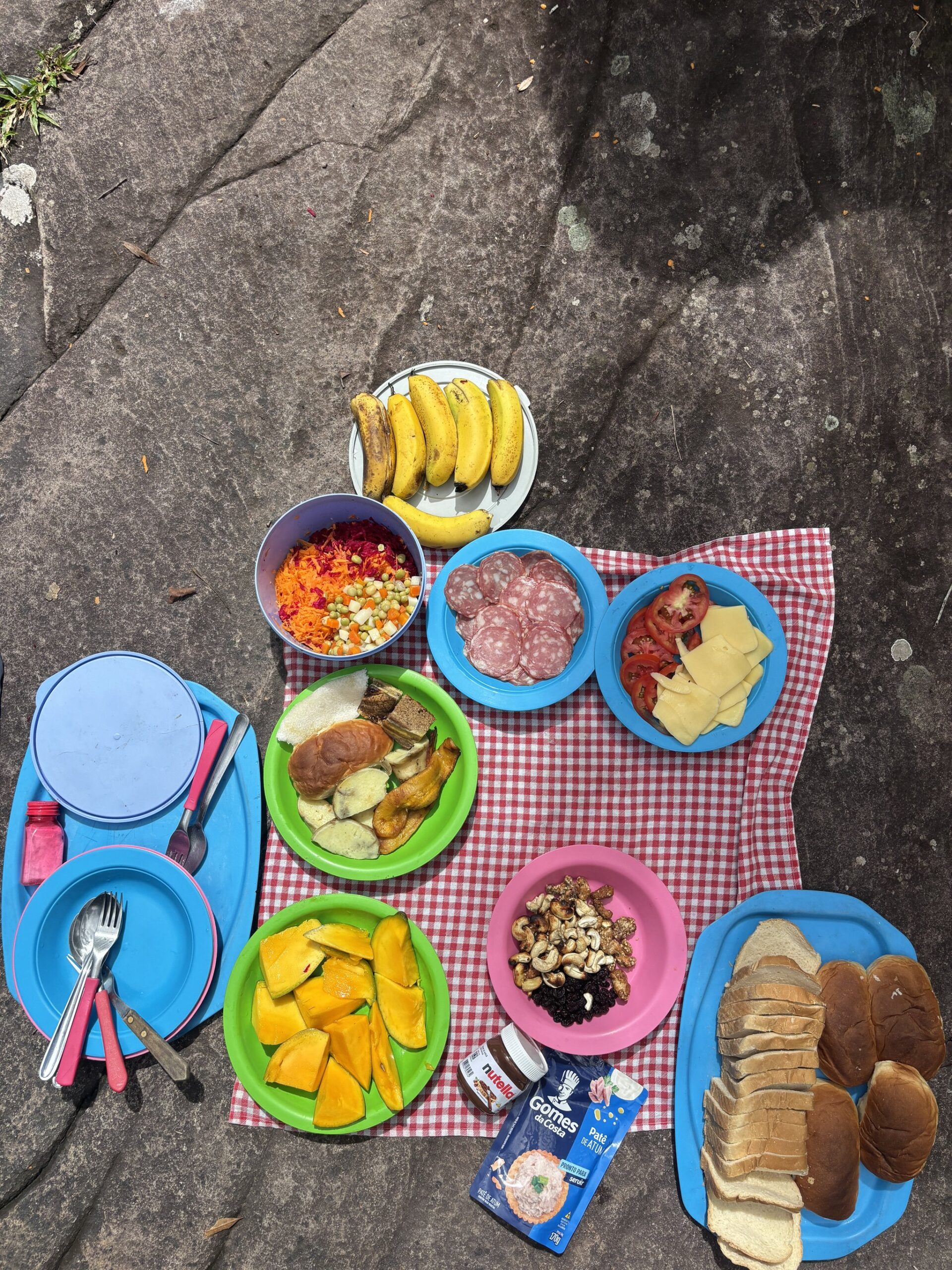 Fuel for the trail — a vibrant spread of fresh fruit, cold cuts and bread served alfresco on bare rock. Colourful plates of fruit, salami, cheese, grated vegetables, nuts and sliced bread laid out on a red-and-white checked cloth on a rock during a Pati Valley hike picnic lunch