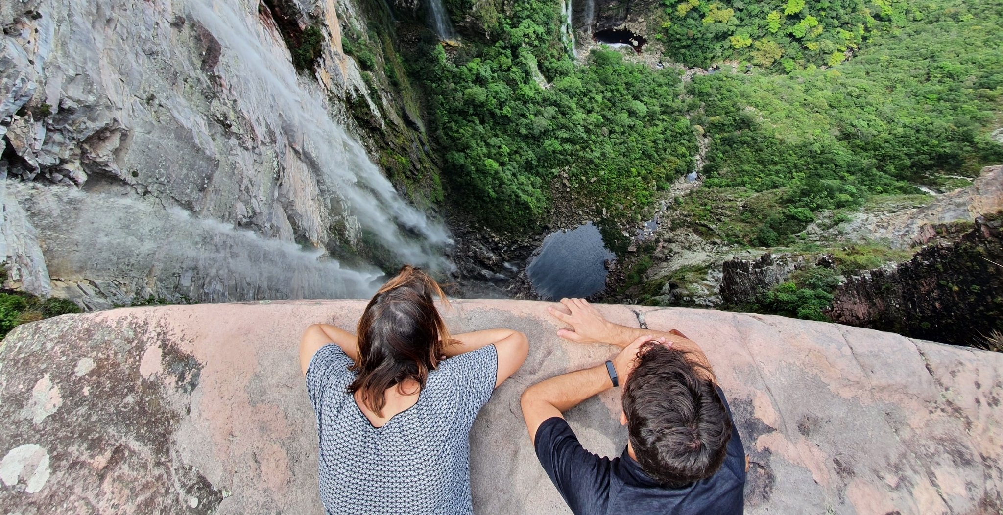 Two trekkers lie on a sandstone ledge peering over the edge at a tall waterfall plunging into a dark pool far below, surrounded by lush green canyon vegetation on the Pati Valley hike