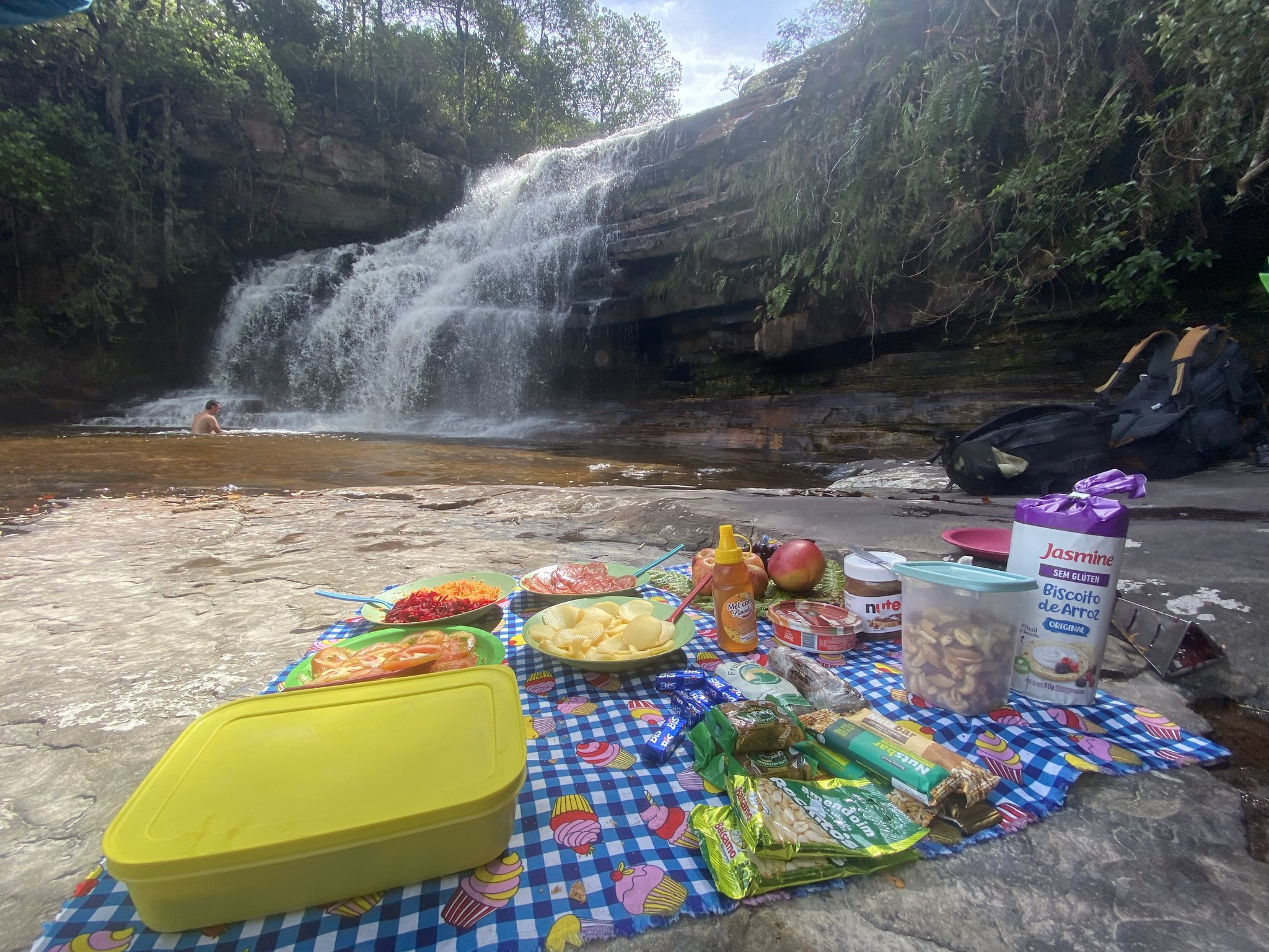 Lunch with a waterfall soundtrack — earned kilometre by kilometre. A colourful trail picnic spread on a checkered blanket beside a sandstone waterfall on the Pati Valley hike, with a swimmer cooling off in the natural pool beyond