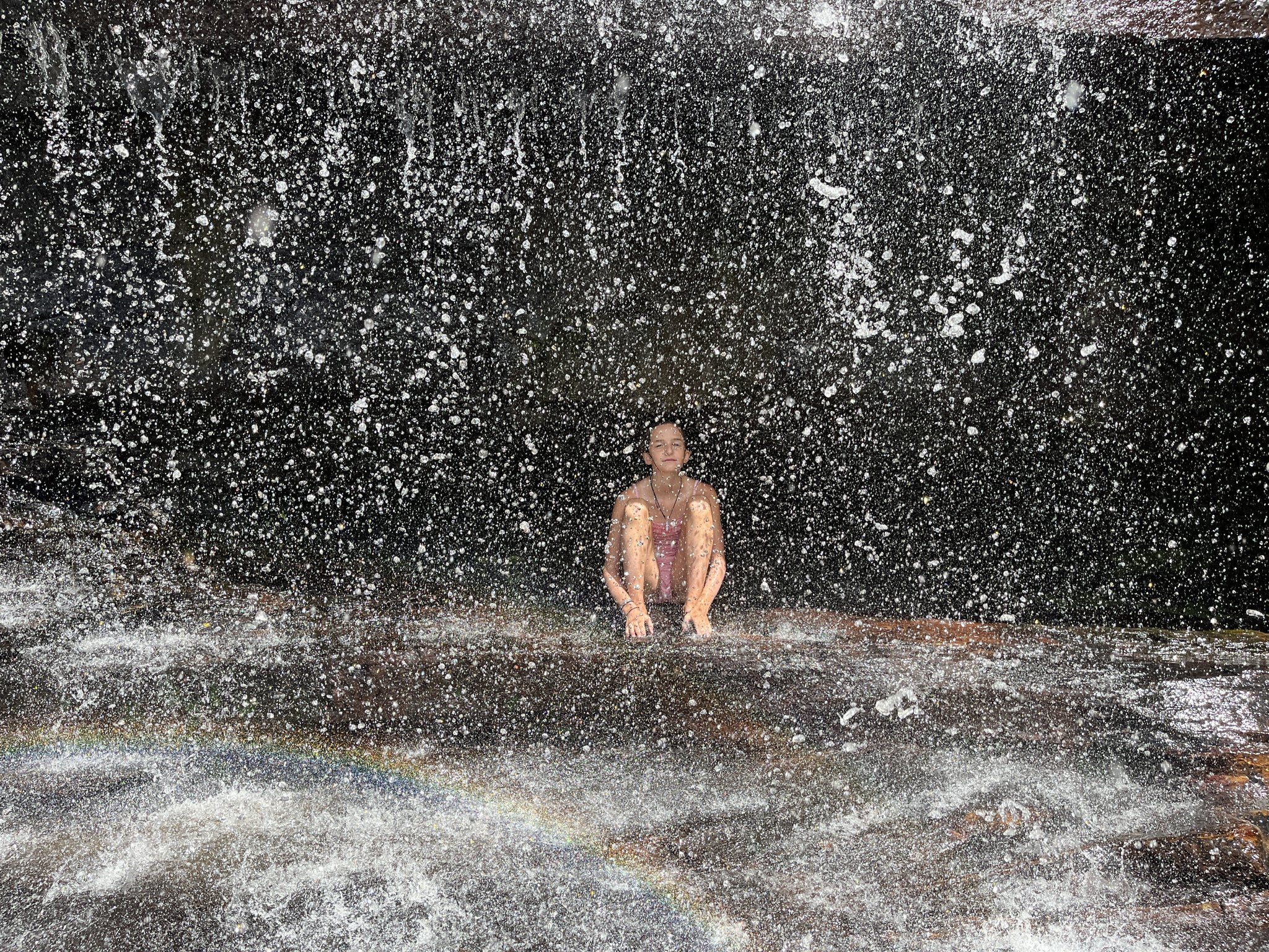 A traveller sits eyes closed on a wet rock ledge beneath a cascading waterfall on the Pati Valley hike, with droplets filling the air and a small rainbow forming at the base