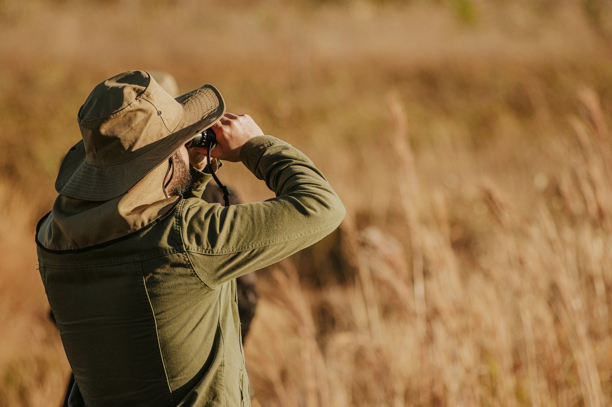 A bearded traveller in a wide-brim hat raises binoculars to scan golden Cerrado grassland during a birdwatching excursion at Pousada Trijunção