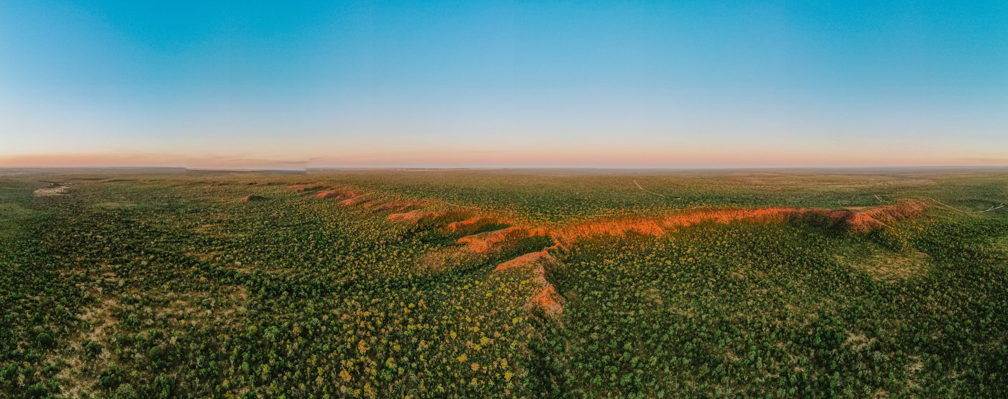 Aerial panoramic view of Pousada Trijunção's vast Cerrado landscape at sunset, with a vivid red laterite ridge cutting through dense green savanna under a pastel pink and blue sky
