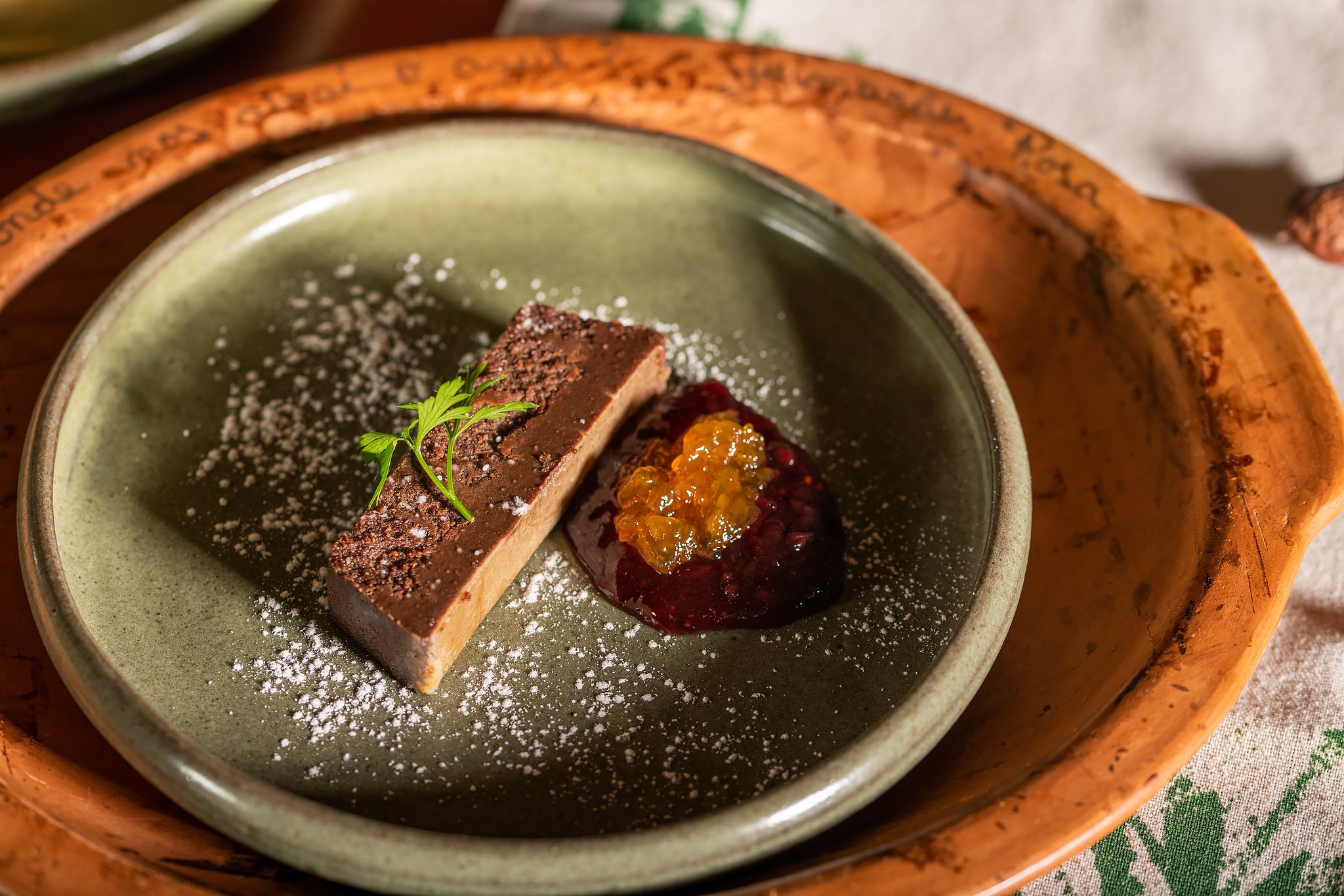 Plated chocolate terrine slice dusted with cocoa powder and icing sugar, served alongside a jewel-toned fruit compote with golden caviar pearls on a sage-green ceramic plate at Pousada Trijunção