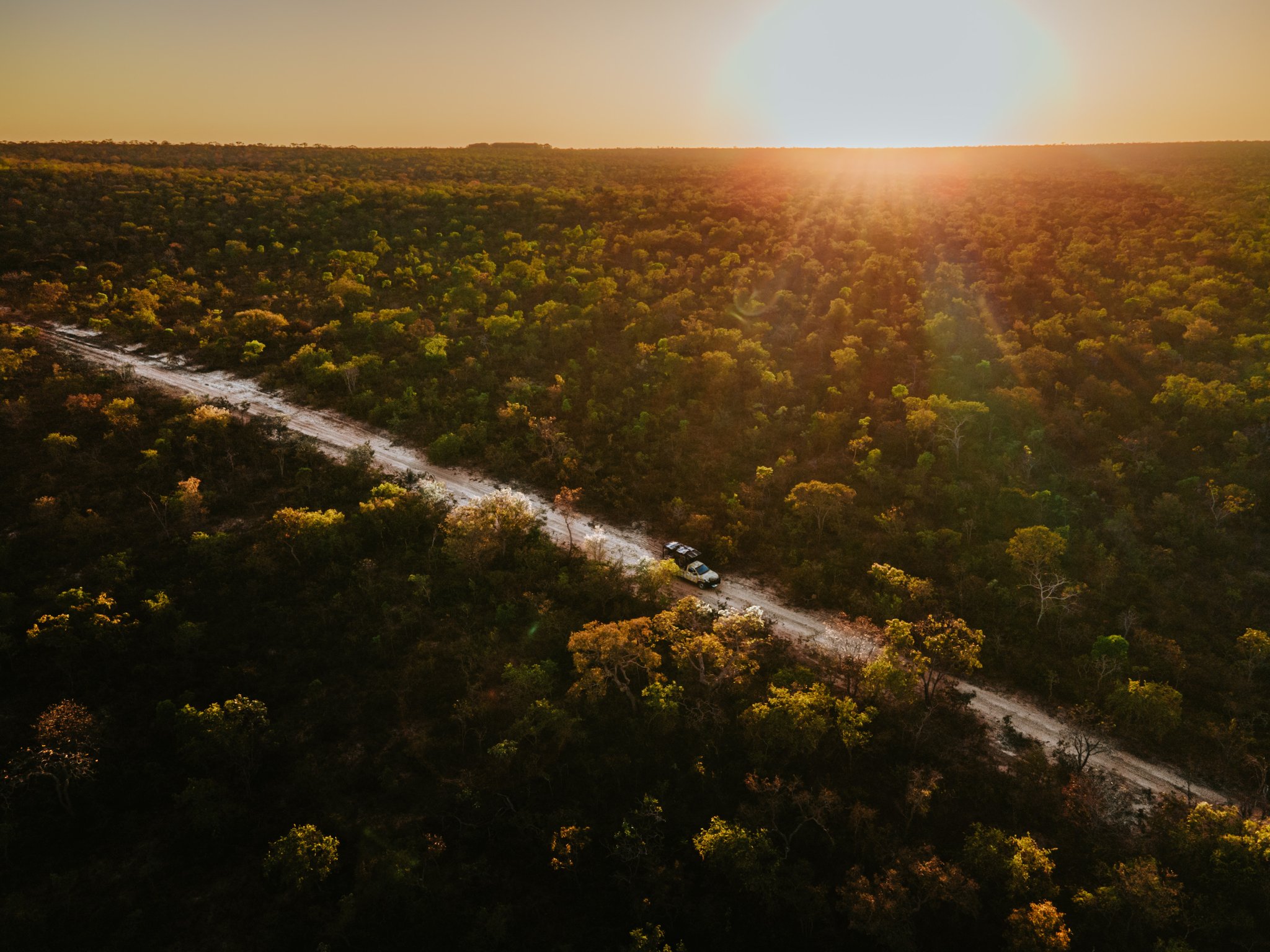 Aerial view of two 4x4 vehicles on a sandy dirt road cutting through dense Cerrado woodland at Pousada Trijunção, bathed in golden sunset light