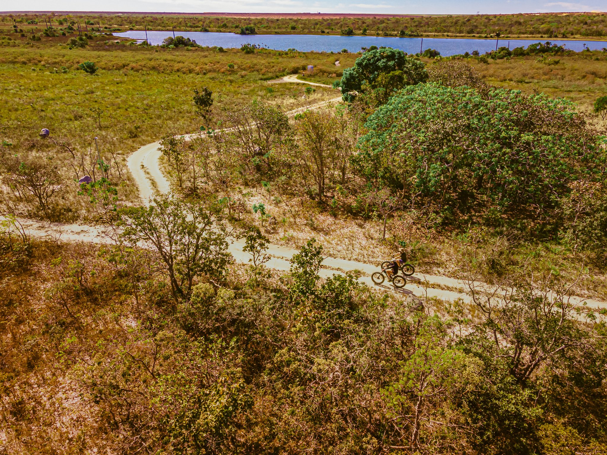 Aerial view of two cyclists riding fat bikes along a winding dirt trail through Cerrado scrubland at Pousada Trijunção, with a blue lagoon visible in the background