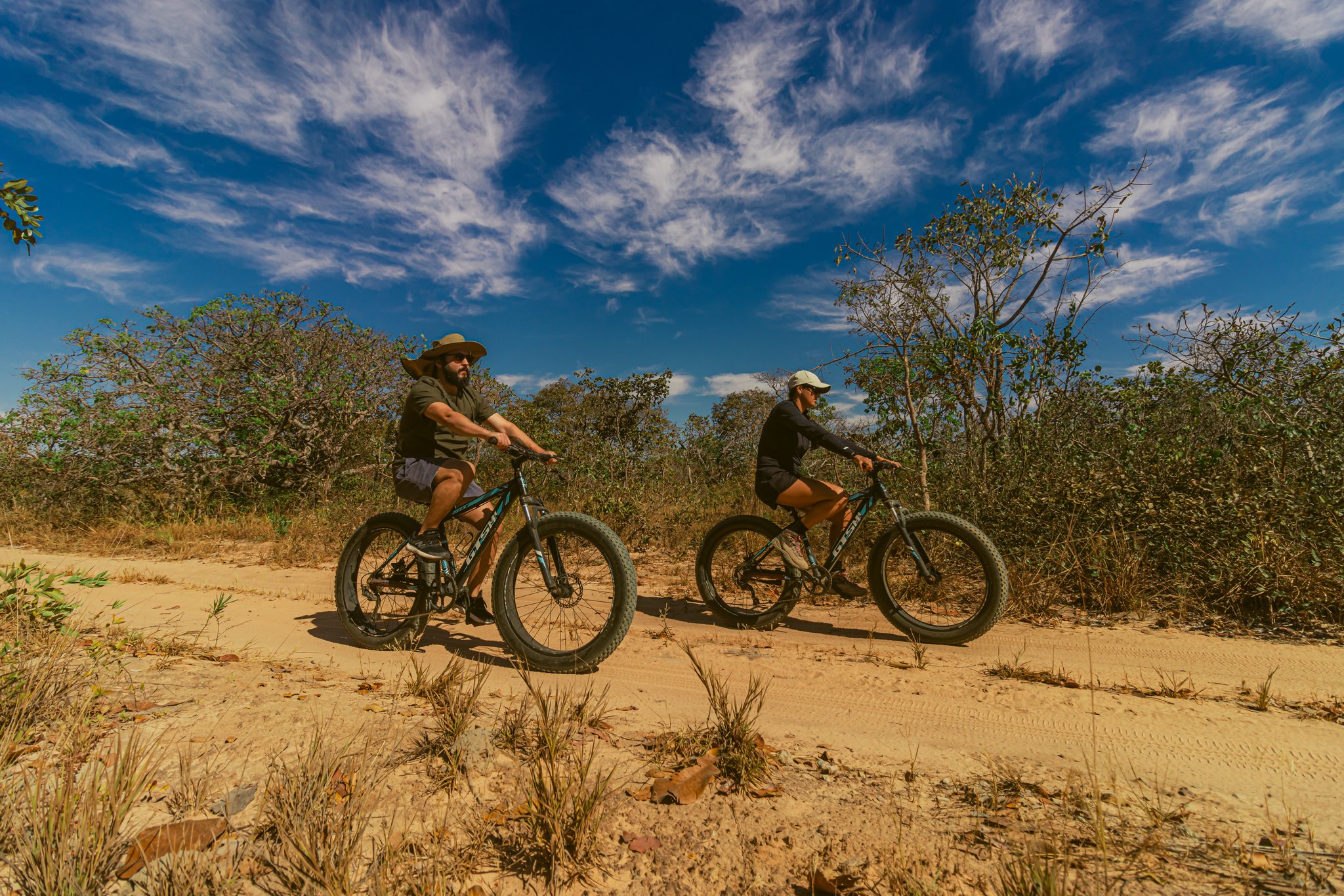 Two cyclists riding fat bikes along a sandy Cerrado trail at Pousada Trijunção, surrounded by native cerrado shrubs under a vivid blue sky