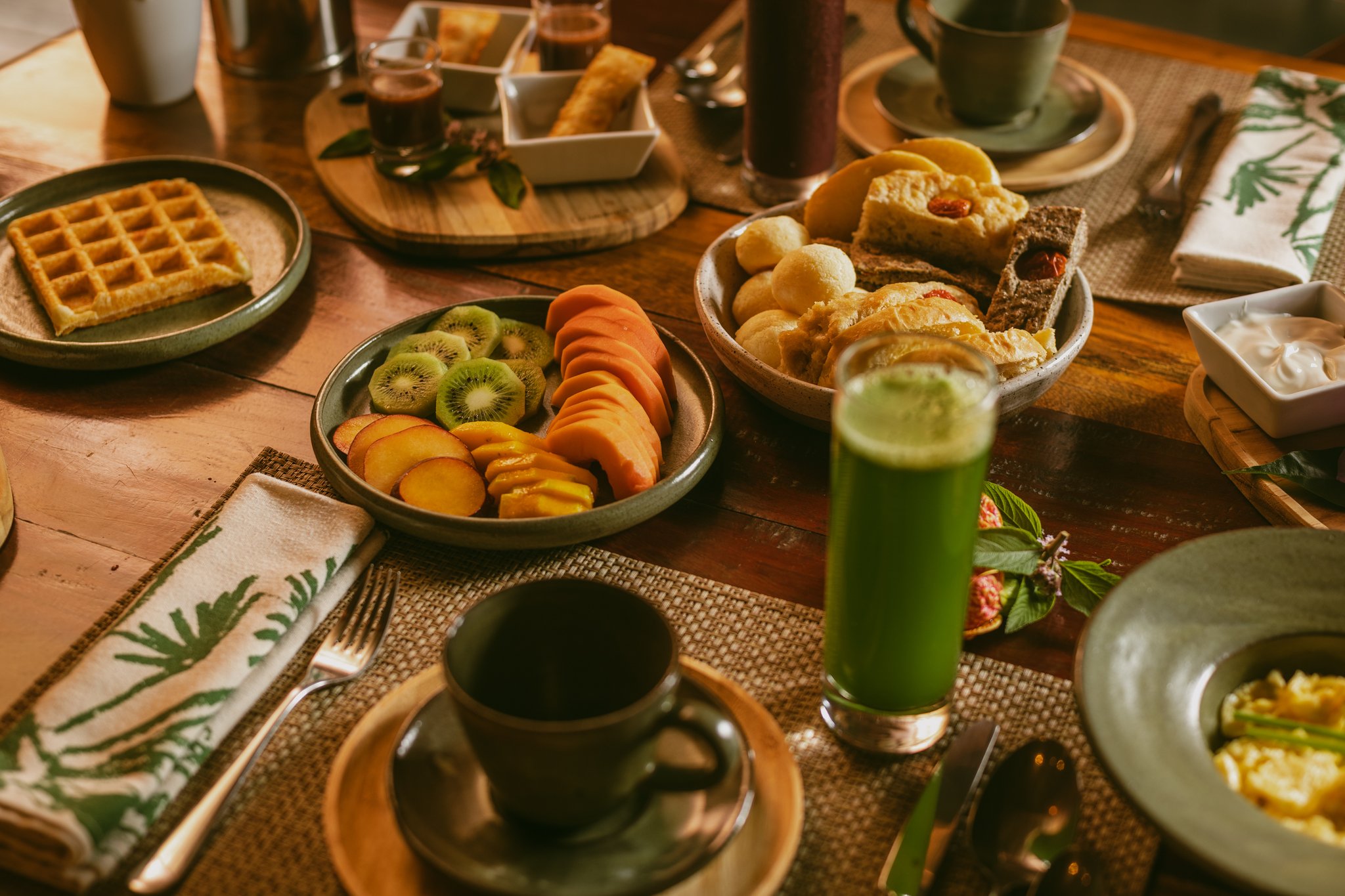 A generous Pousada Trijunção breakfast table laid with sliced kiwi, peach and papaya, golden waffles, warm bread rolls, a vibrant green juice and a ceramic coffee mug on a woven placemat