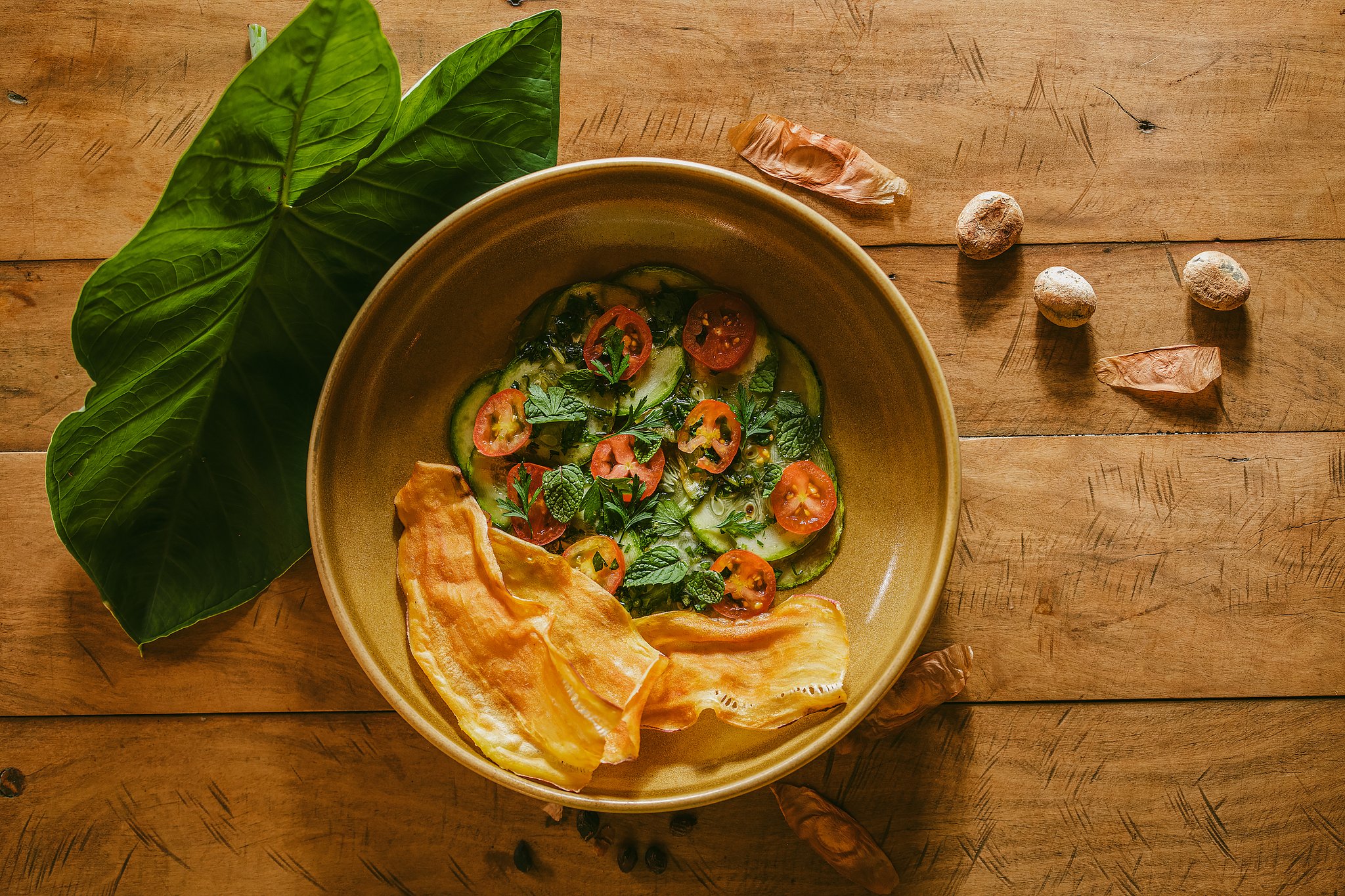 Overhead view of a ceramic bowl filled with sliced zucchini, cherry tomatoes and fresh herbs served with crispy chips at Pousada Trijunção, surrounded by tropical leaves and wild seeds on a rustic wooden table