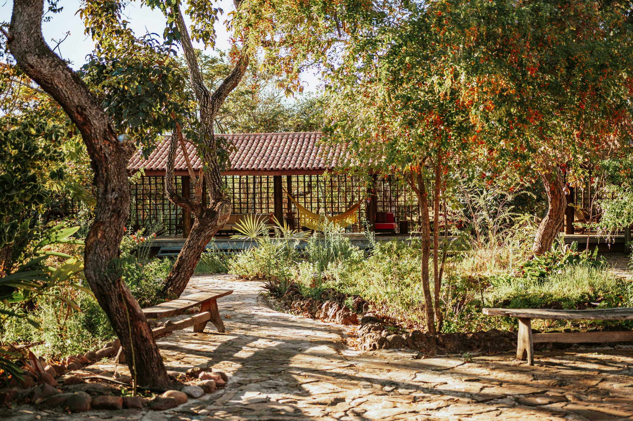 Sun-dappled stone pathway at Pousada Trijunção leading through a lush garden of native Cerrado trees toward an open-air pavilion with a hammock and terracotta-tiled roof
