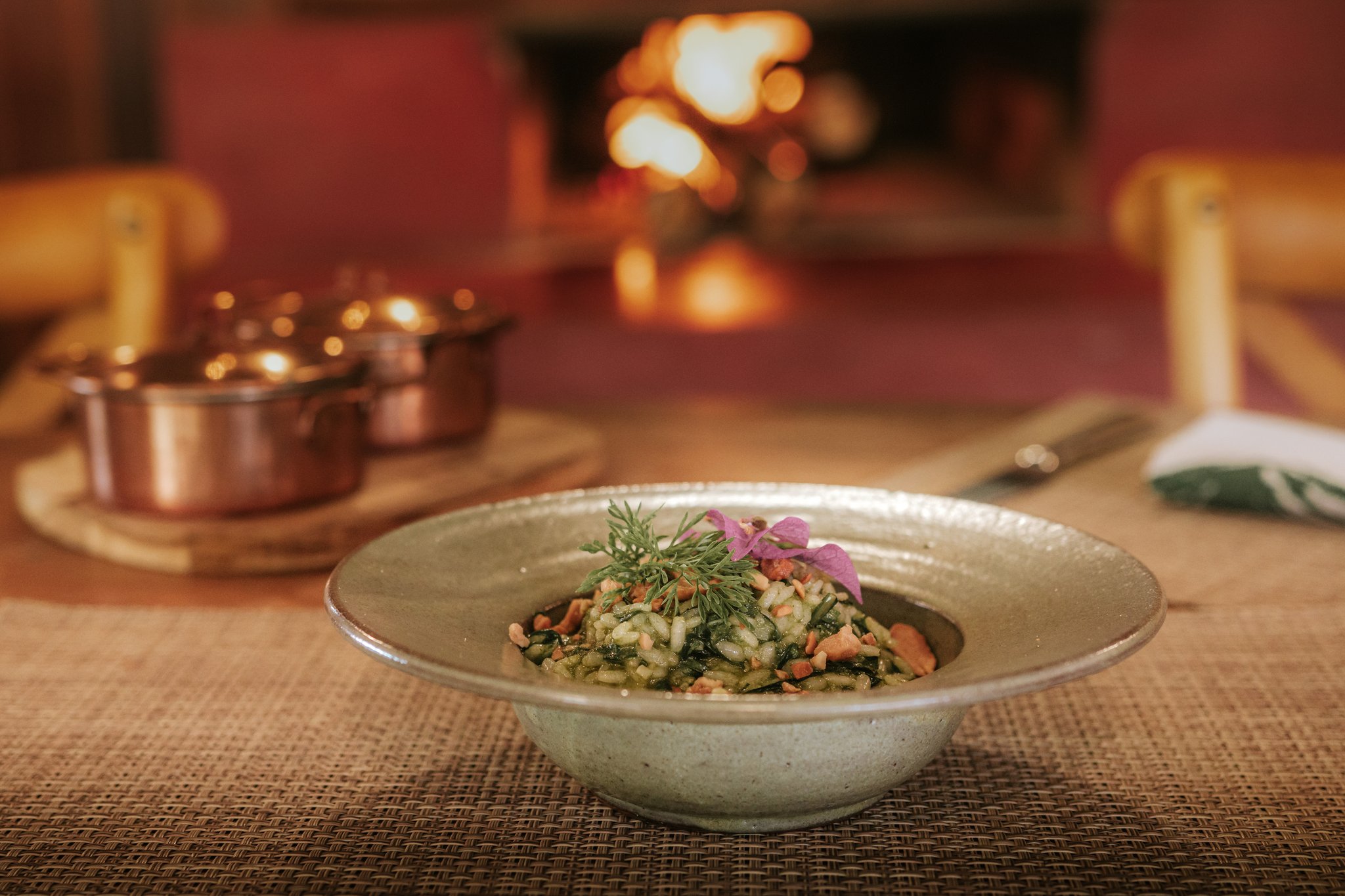 A handcrafted ceramic bowl of herb risotto garnished with fresh dill and a pink edible flower, served fireside at Pousada Trijunção with copper pots glowing in the warm background
